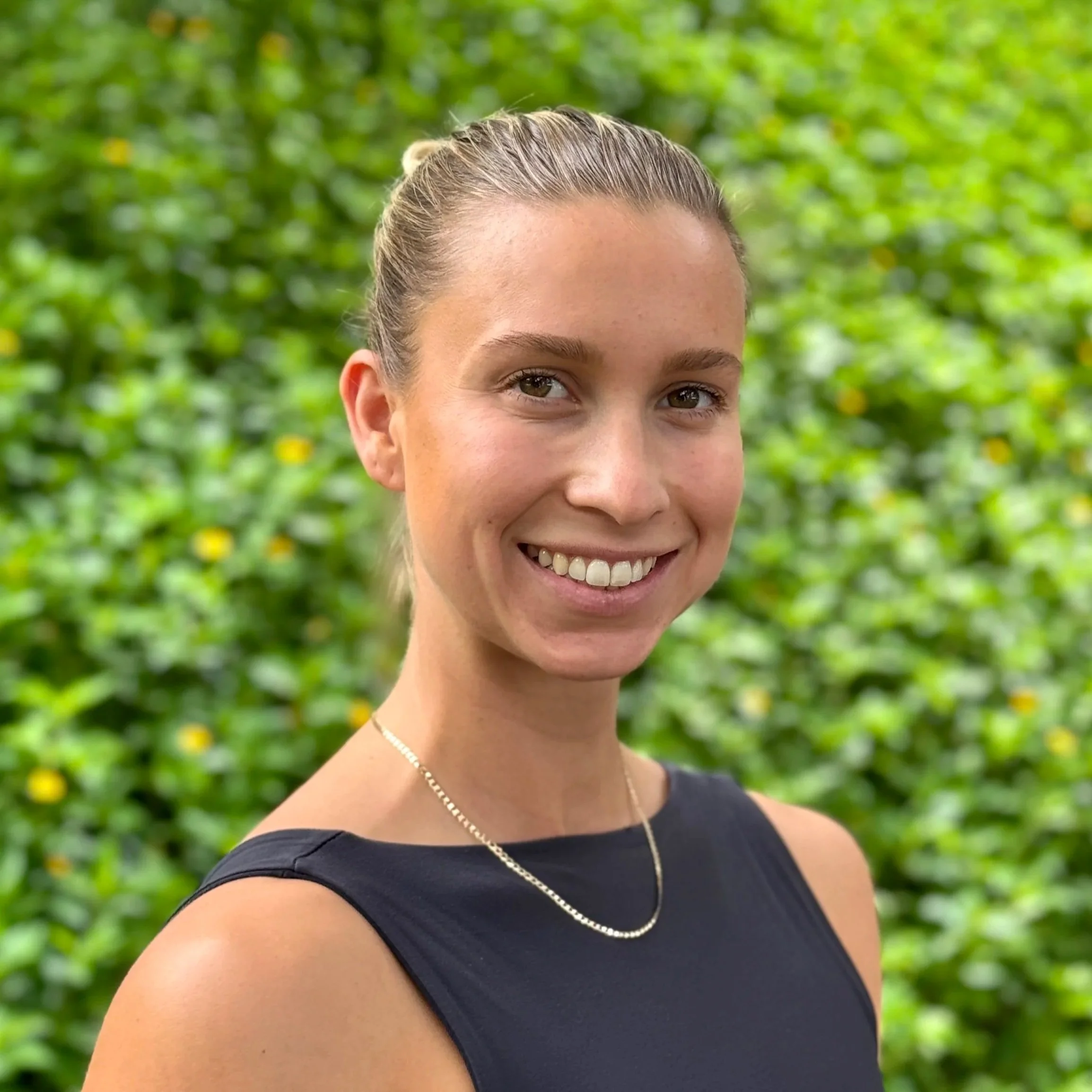 Close-up of a smiling woman with light skin, brown hair pulled back in a bun, wearing a black top and a pearl necklace, standing outdoors with green foliage in the background.