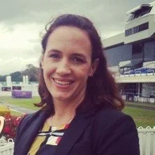 A woman with dark hair smiling outdoors at a racetrack or sports venue, with buildings and a cloudy sky in the background.