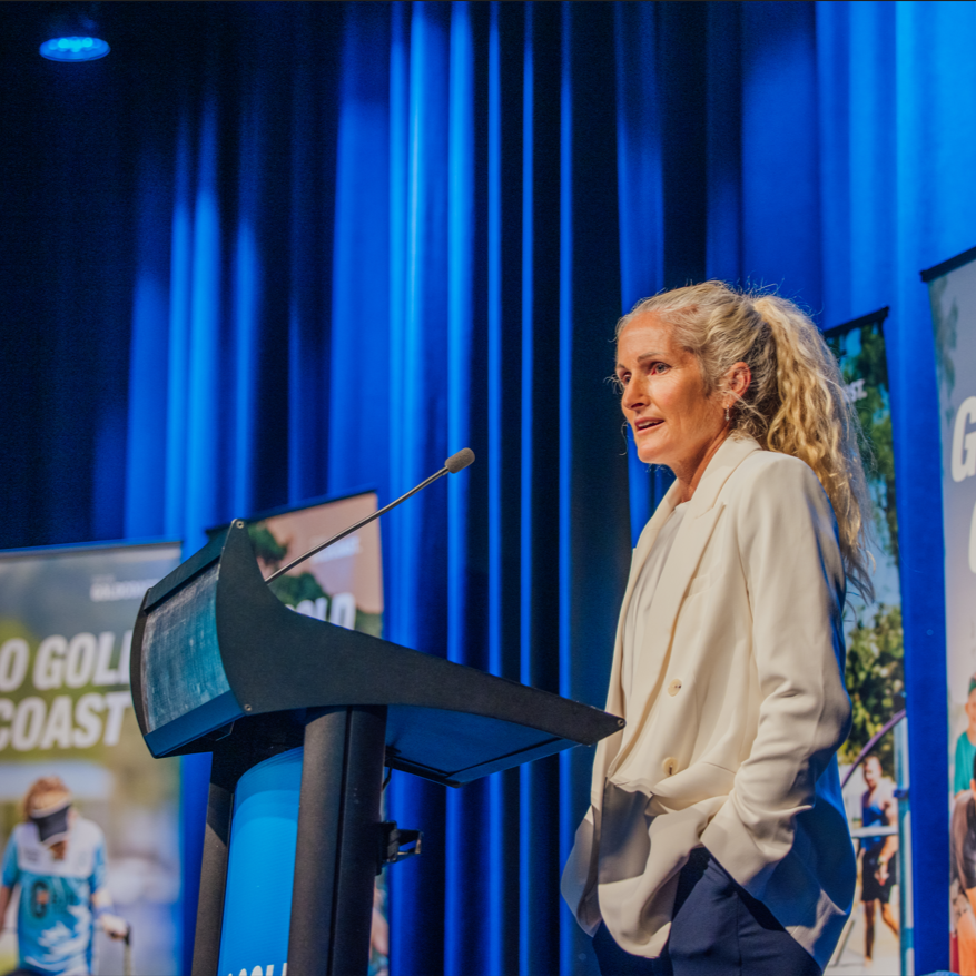 A woman with blonde curly hair tied in a ponytail, wearing a white blazer, speaking behind a podium on a stage with blue curtains and banners in the background.