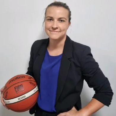 A woman in a blazer and blue shirt holding a basketball, standing against a plain white background.