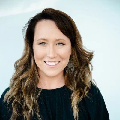 A woman with long, wavy brown hair and blue eyes, smiling in front of a light blue background.
