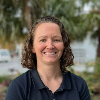 A smiling woman with curly brown hair wearing a navy blue collared shirt outdoors, with trees and a blurred background.