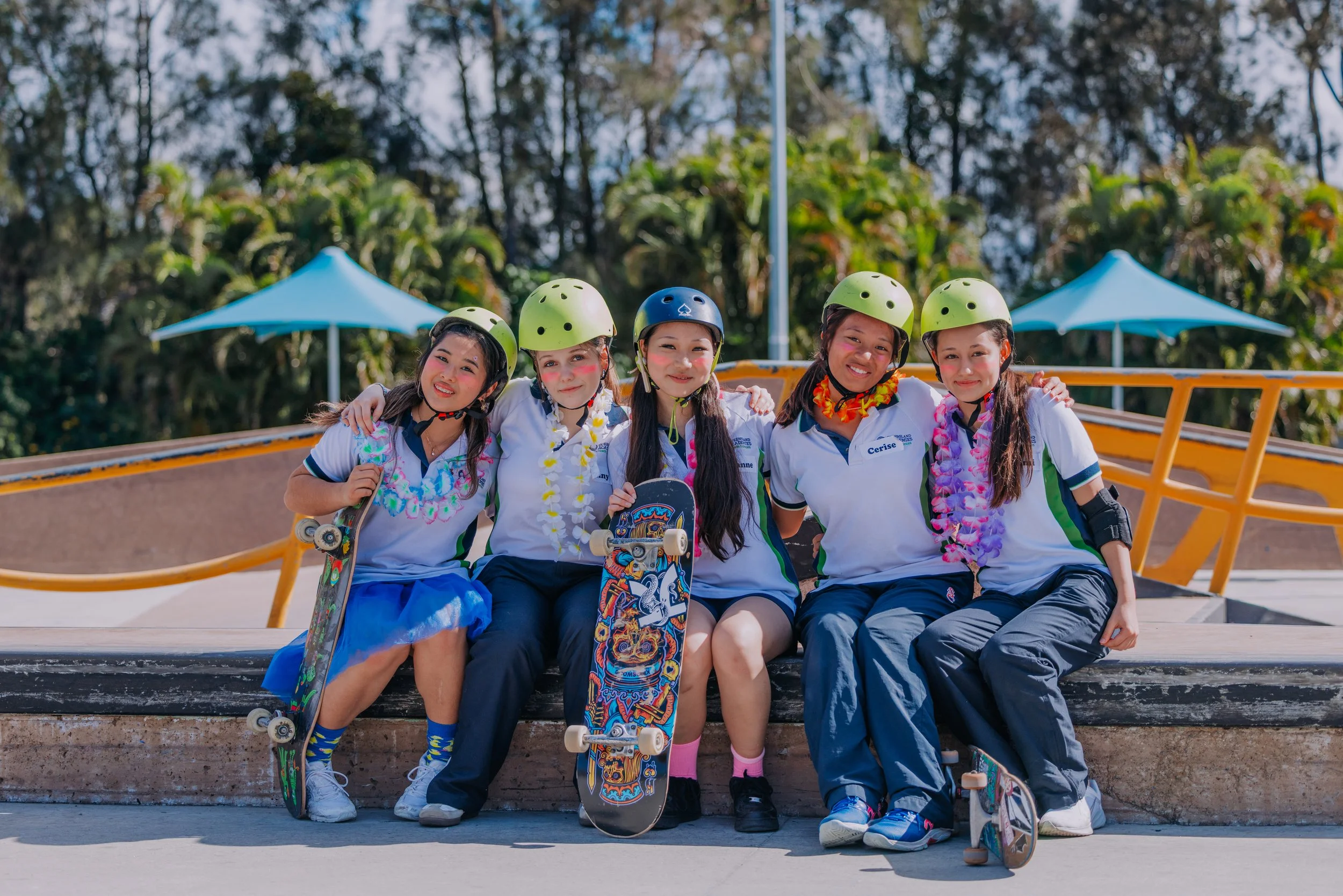 Five young girls wearing helmets and casual clothes, sitting together with skateboards, smiling outdoors on a sunny day with trees and blue umbrellas in the background.