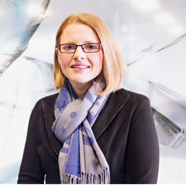A woman with red hair, glasses, wearing a dark blazer and a blue polka dot scarf, smiling in front of a blurred background.