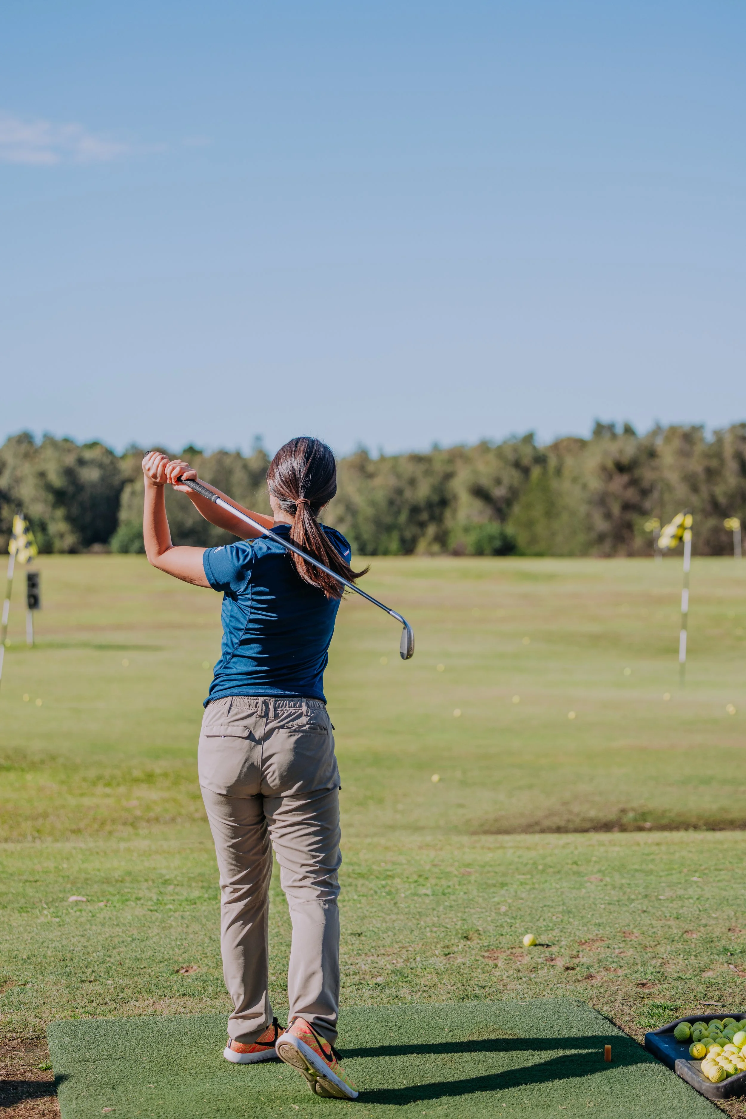 A woman practicing golf at a driving range, standing on a mat with a golf club, with a basket of golf balls nearby, under a blue sky with trees in the background.