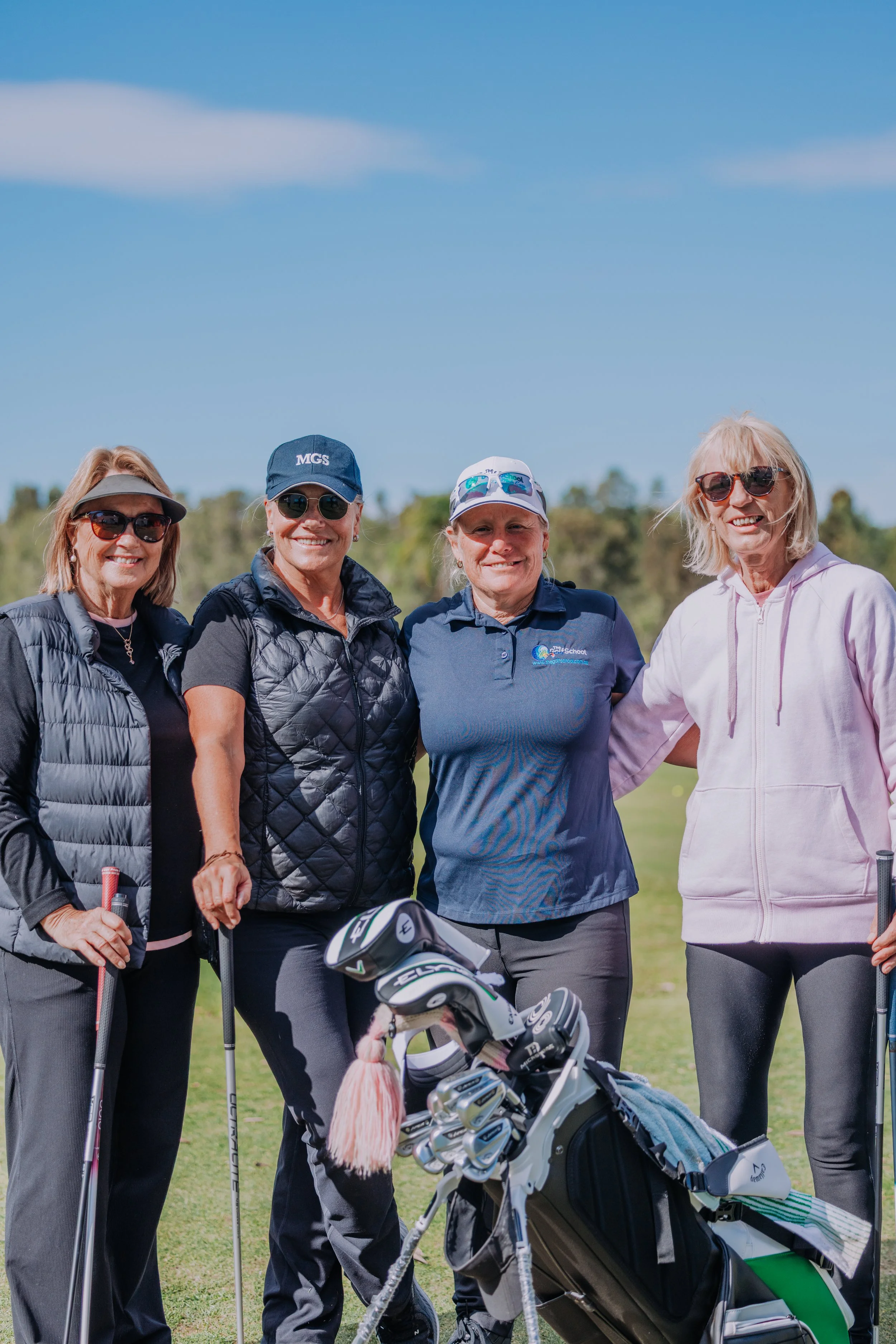 Four women standing on a golf course, smiling and posing for a picture, with golf clubs and a golf bag in the foreground. They are dressed in casual golf attire with hats and sunglasses, and there are trees and a blue sky in the background.