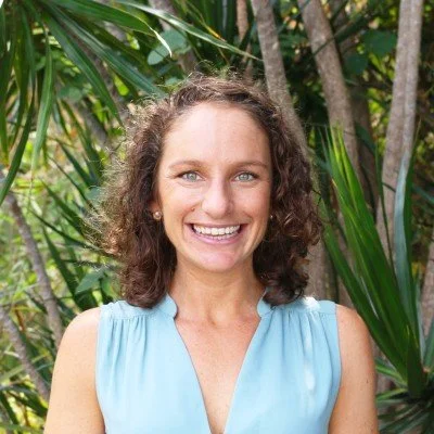 A woman with curly brown hair smiling outdoors among green plants and trees.