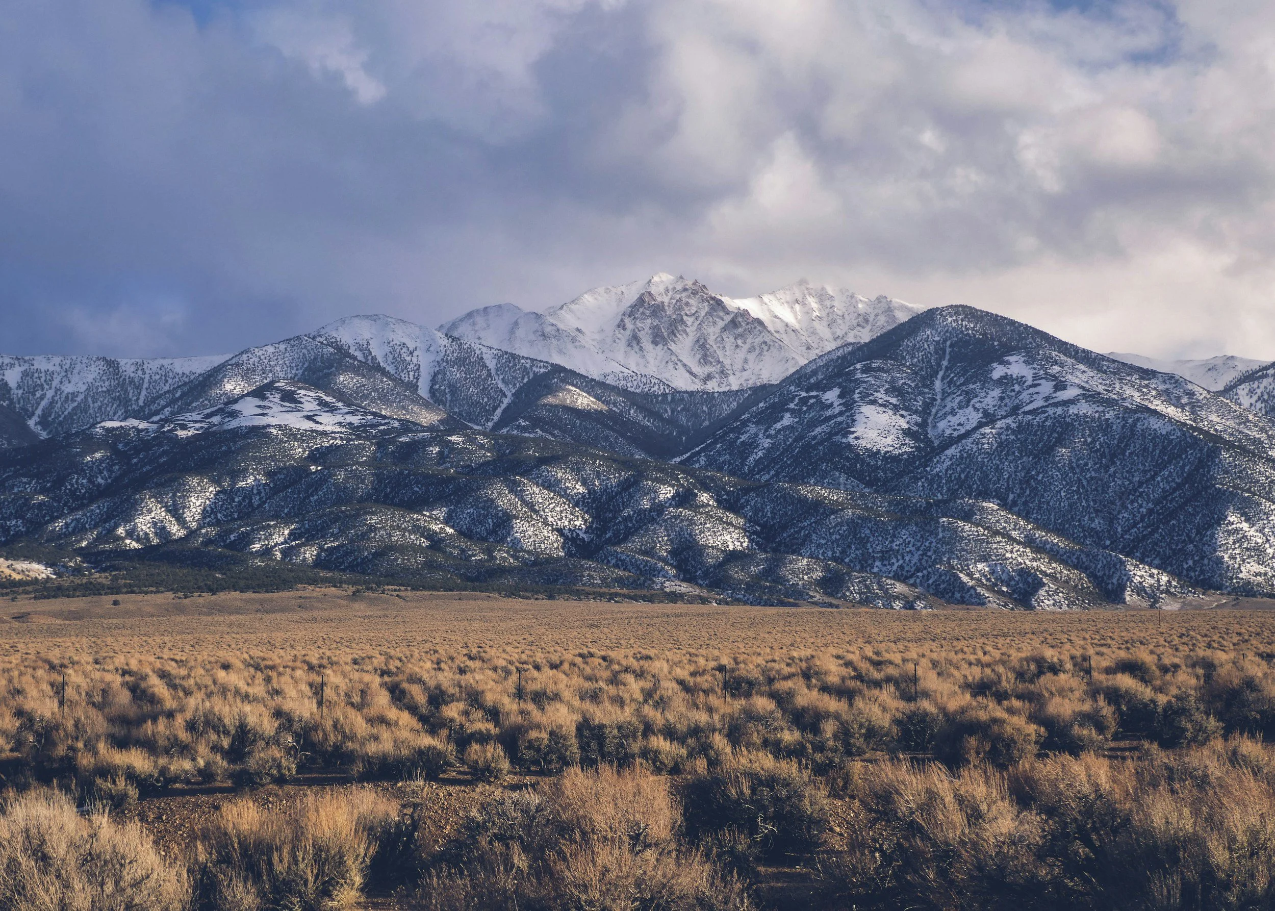 Snow-capped mountains under a cloudy sky with a flat, dry foreground.