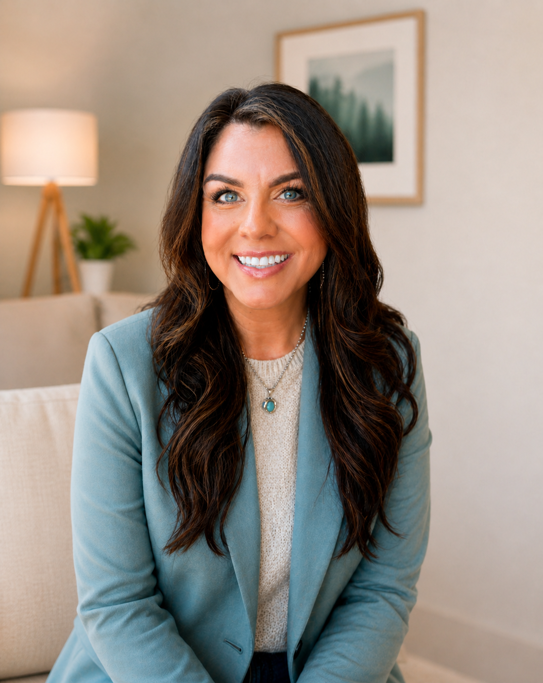 A woman with long, wavy brown hair and blue eyes smiling, sitting in a cozy living room with a lamp and a framed painting in the background.
