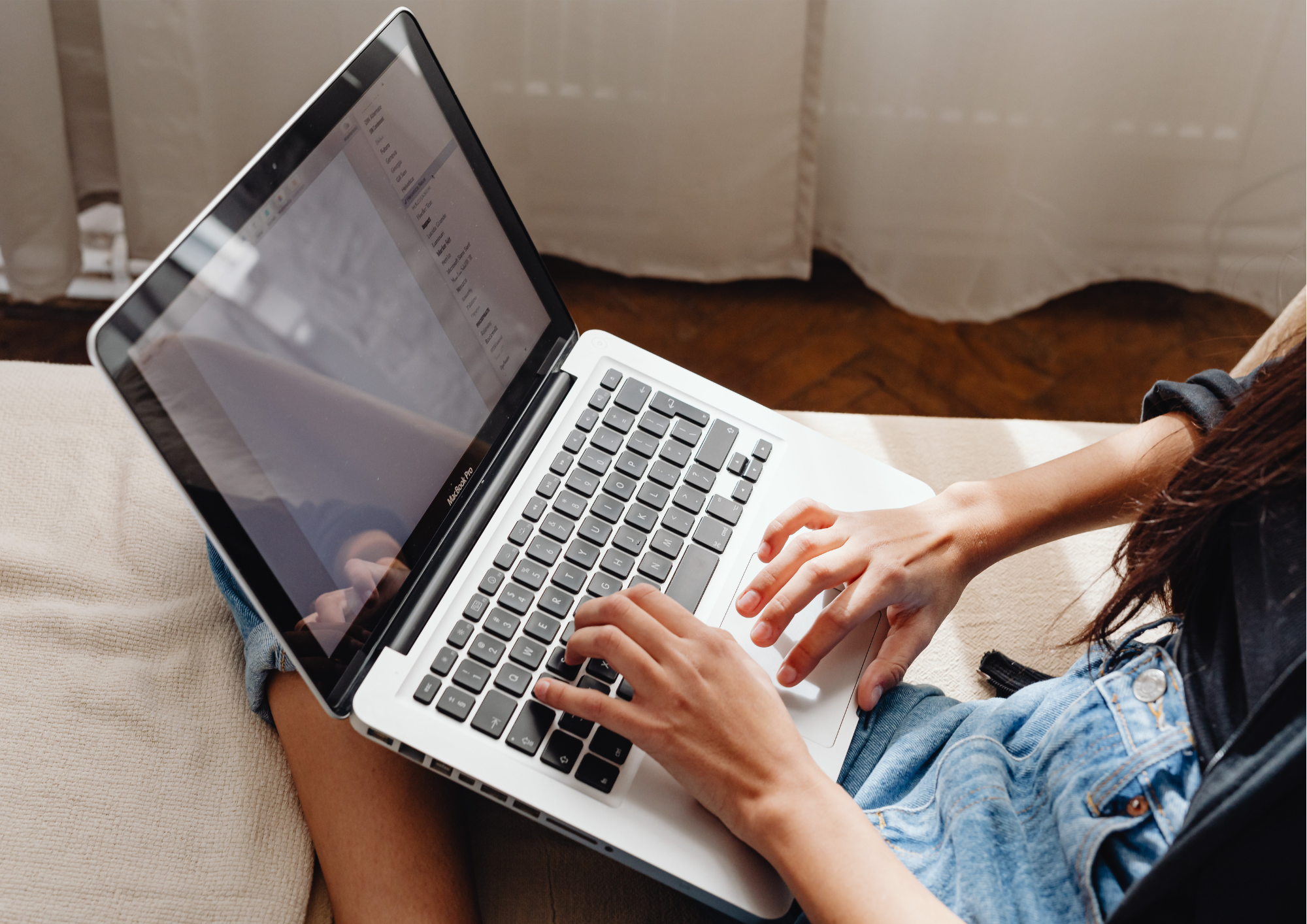 A person wearing a blue denim jacket and black shirt sitting on a beige couch, working on a silver MacBook laptop with a reflective screen showing email inbox.