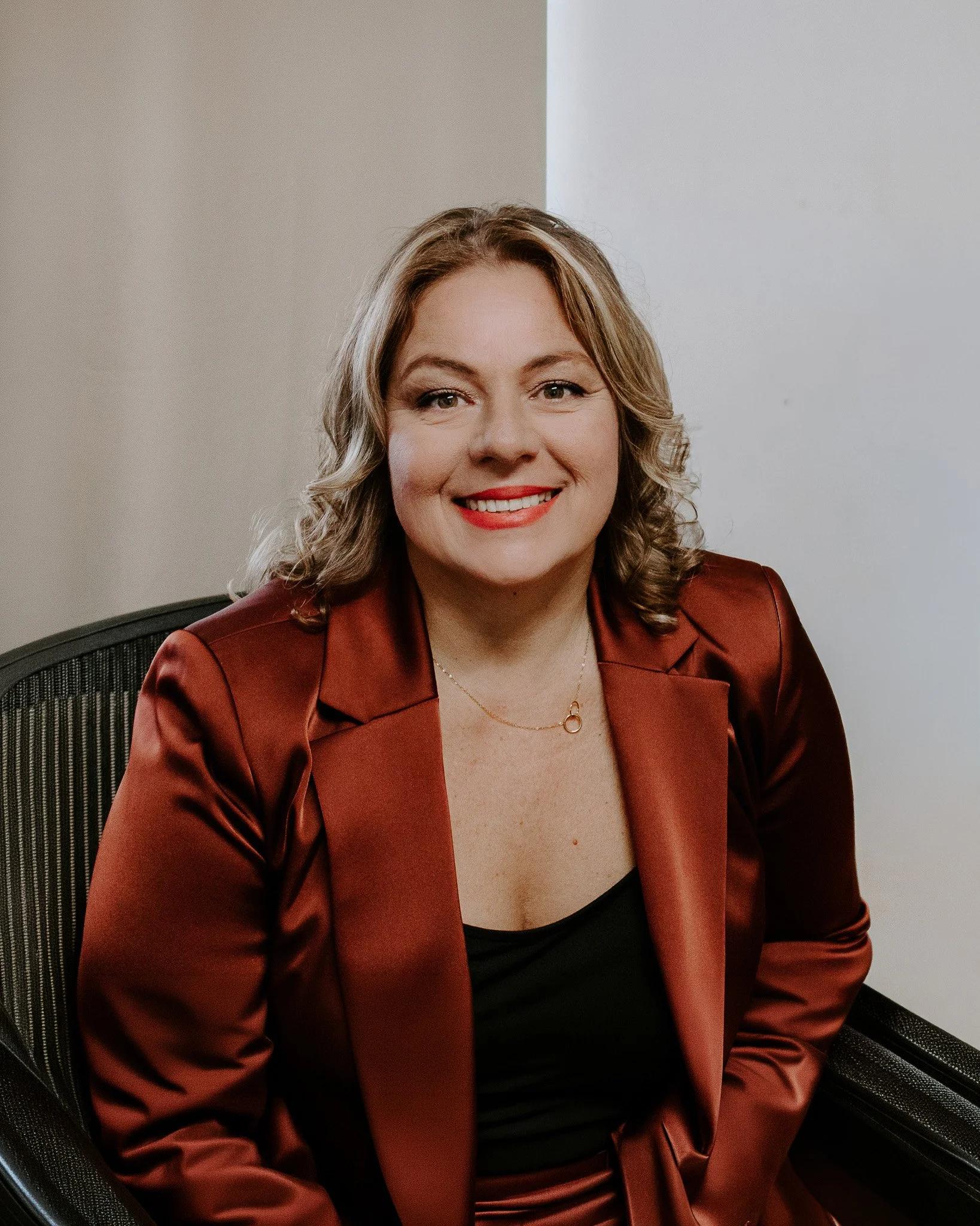 A woman with wavy blonde hair and red lipstick sitting in a black office chair, wearing a fitted black top and a brown blazer. The background features a plain light-colored wall.
