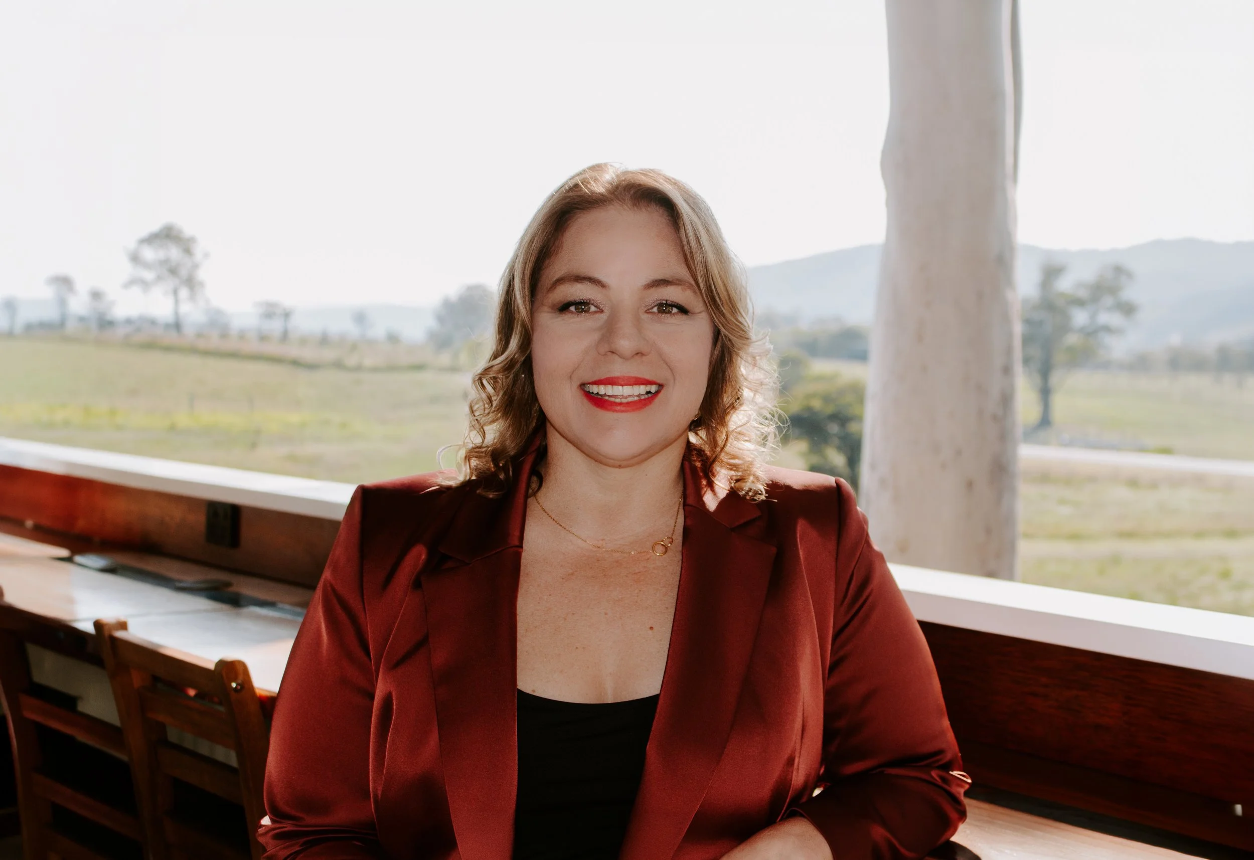 A woman with curly red hair and red lipstick smiling while sitting at a wooden table in a bright room with a window overlooking a scenic outdoor area with trees and hills.