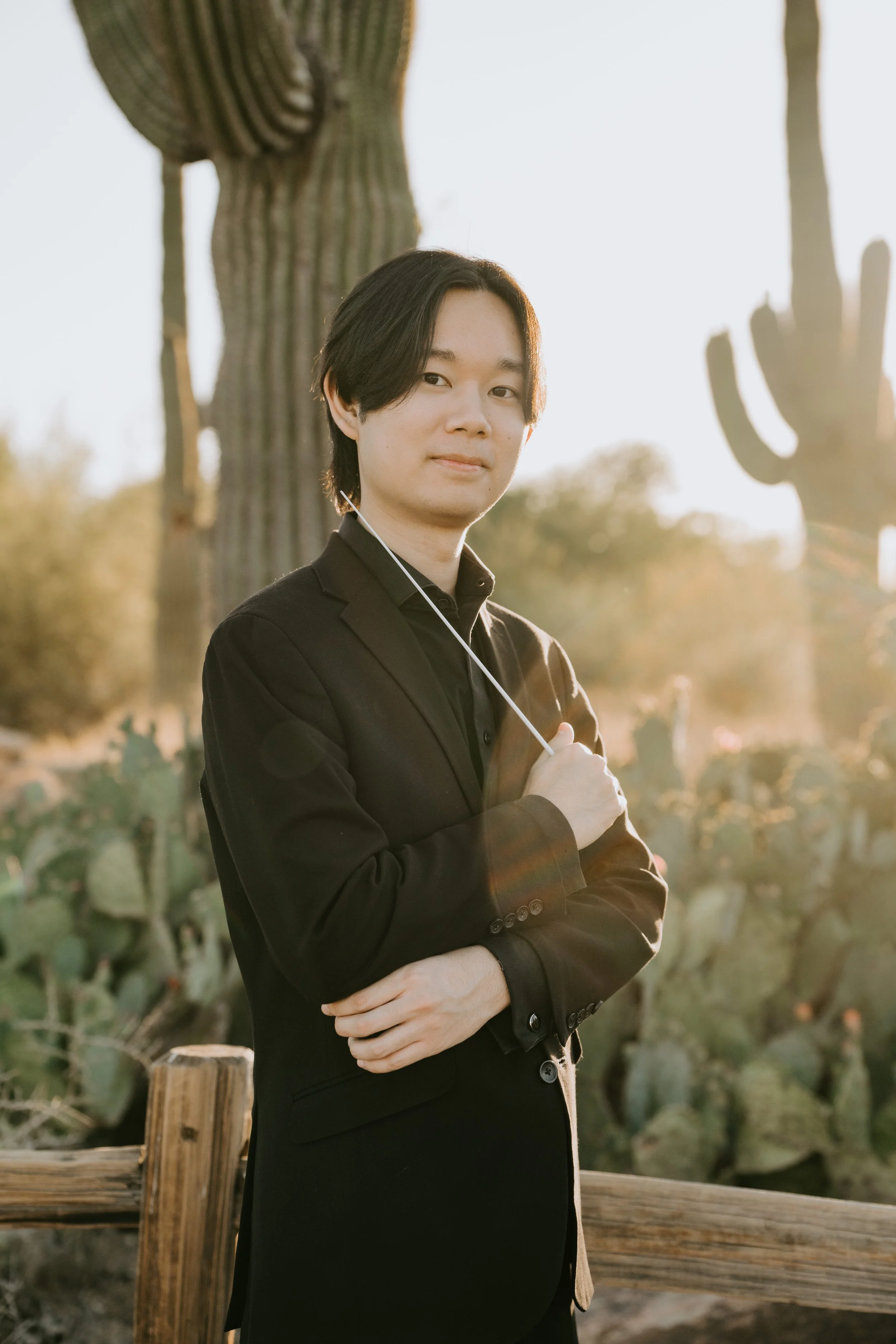 A young man with dark hair wearing a black blazer and shirt, standing outdoors near a wooden fence and large cactus plants during sunset.