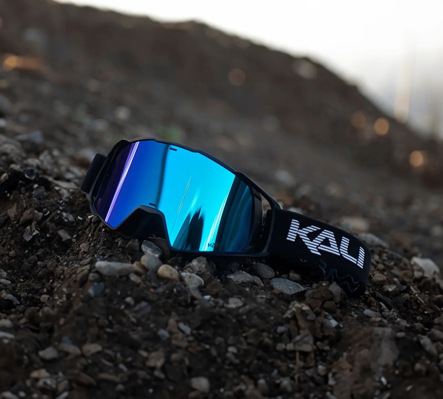 Black sports sunglasses with reflective blue lenses lying on dirt and gravel ground.