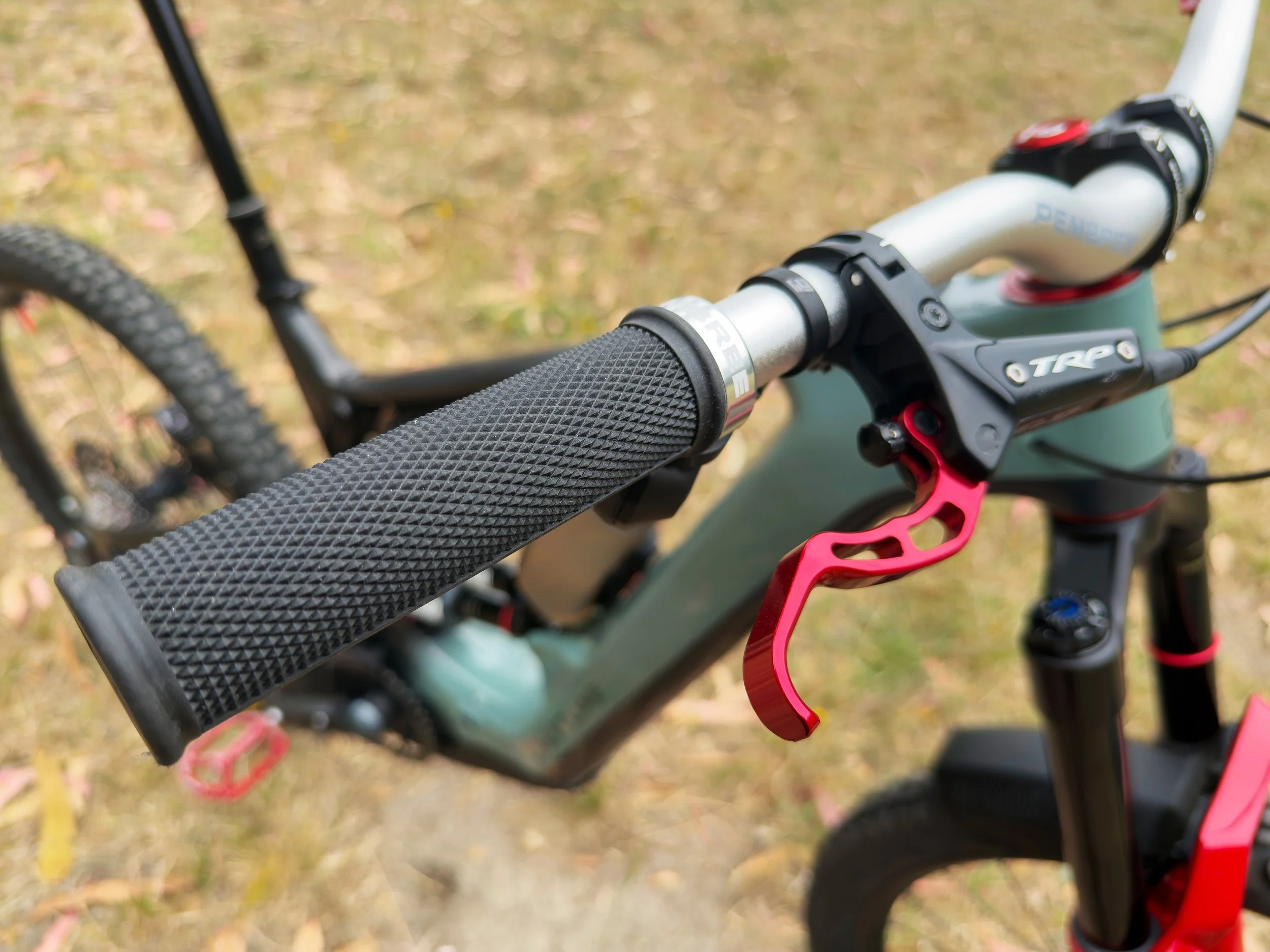 Close-up of a mountain bike handlebar with textured grip, red gear shifter, and black brake lever, set outdoors on grass.