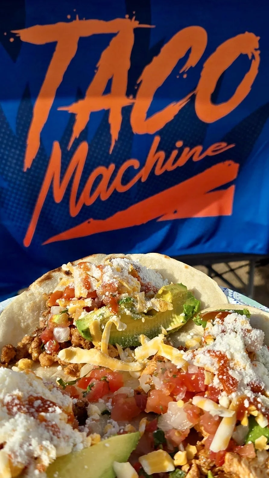 Close-up of a taco with ground meat, chopped tomatoes, cheese, and avocado in a soft tortilla, with a taco shop bag in the background.