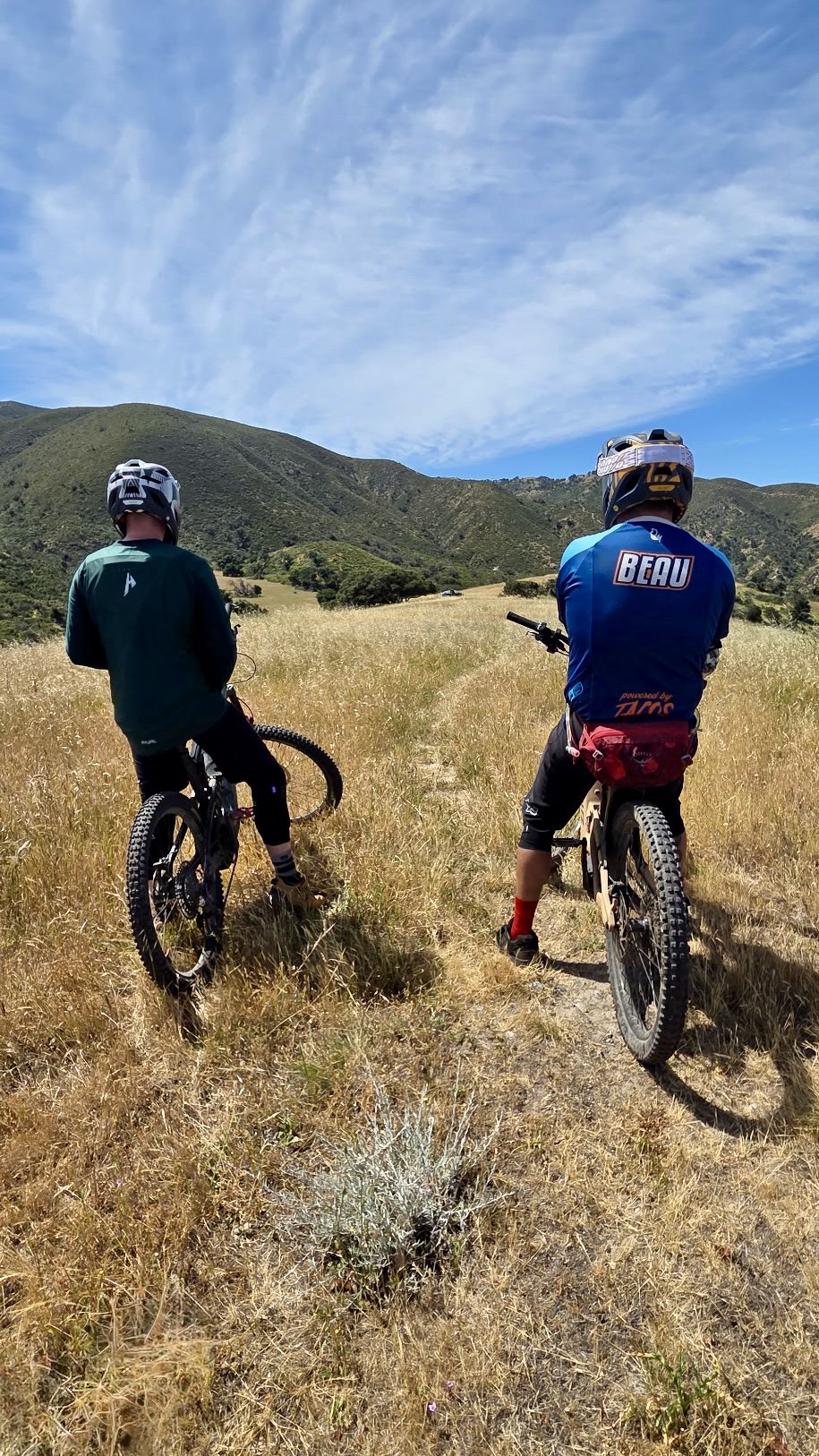 Two mountain bikers with helmets sitting on bikes, looking toward scenic rolling hills and blue sky.