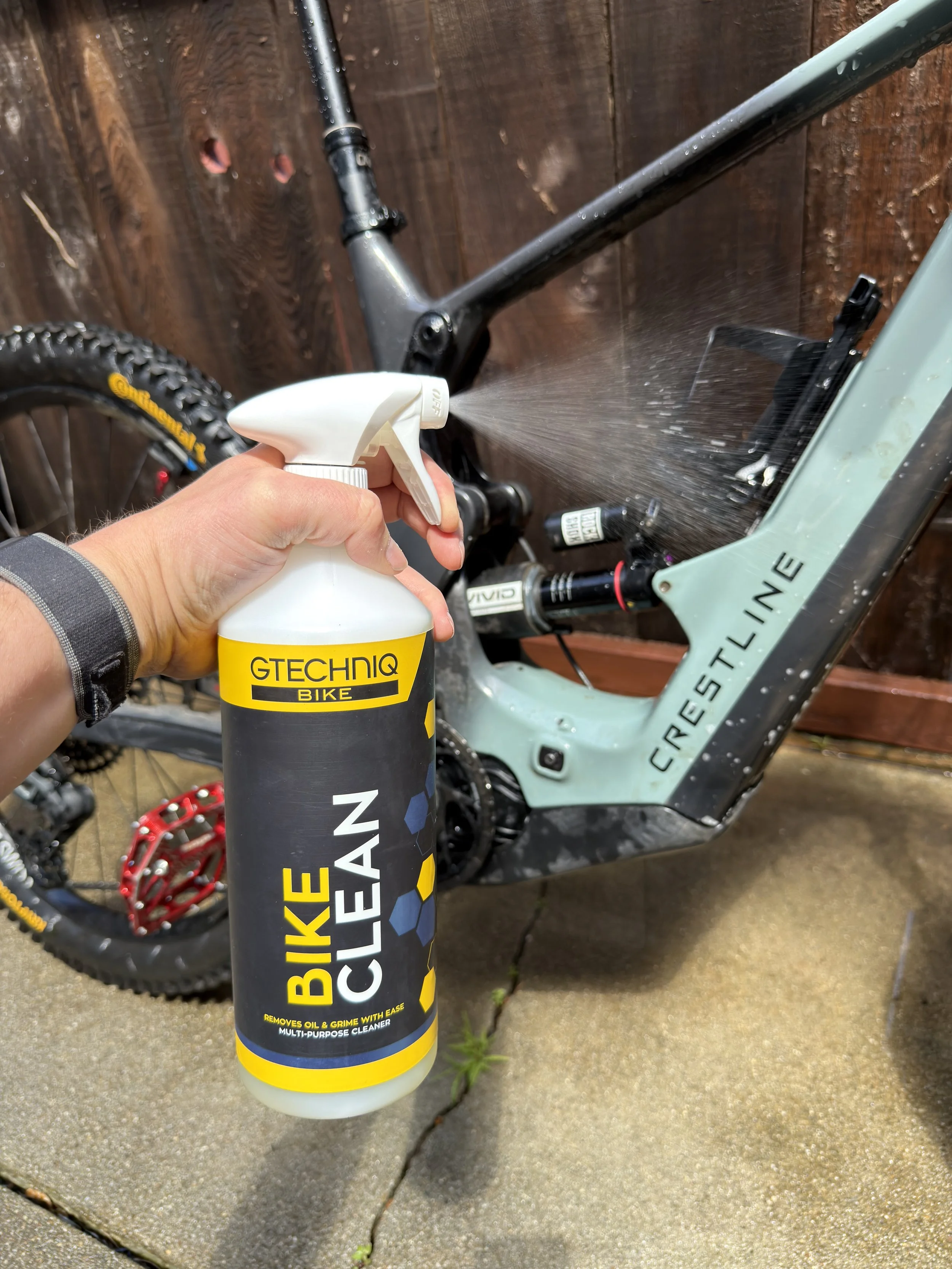 Person holding a yellow and black can of bike cleaner spraying the frame of a light-colored mountain bike.