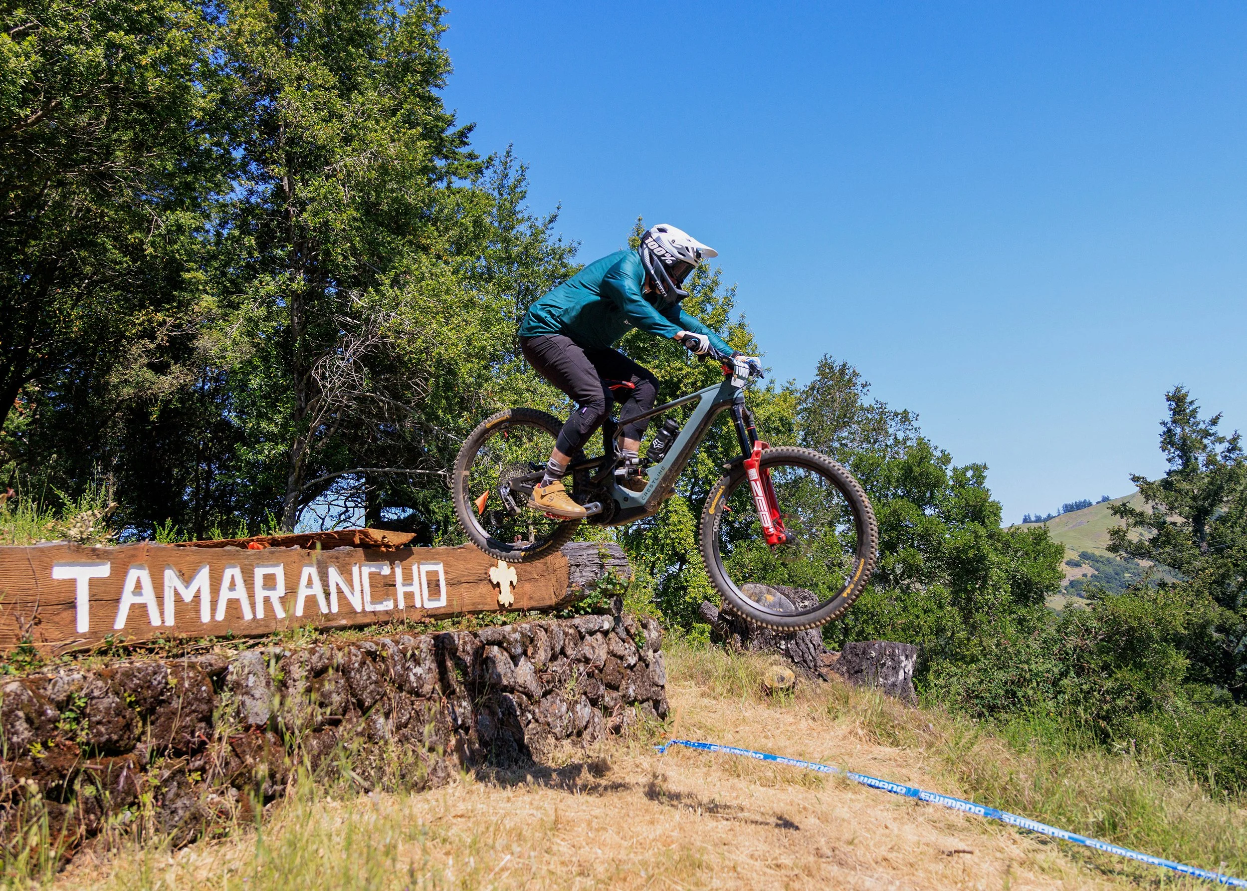 A mountain biker in a teal jersey, black pants, and a white helmet riding a bike over a dirt log obstacle labeled 'Tamaranch' in a forested outdoor area with trees and clear blue sky.