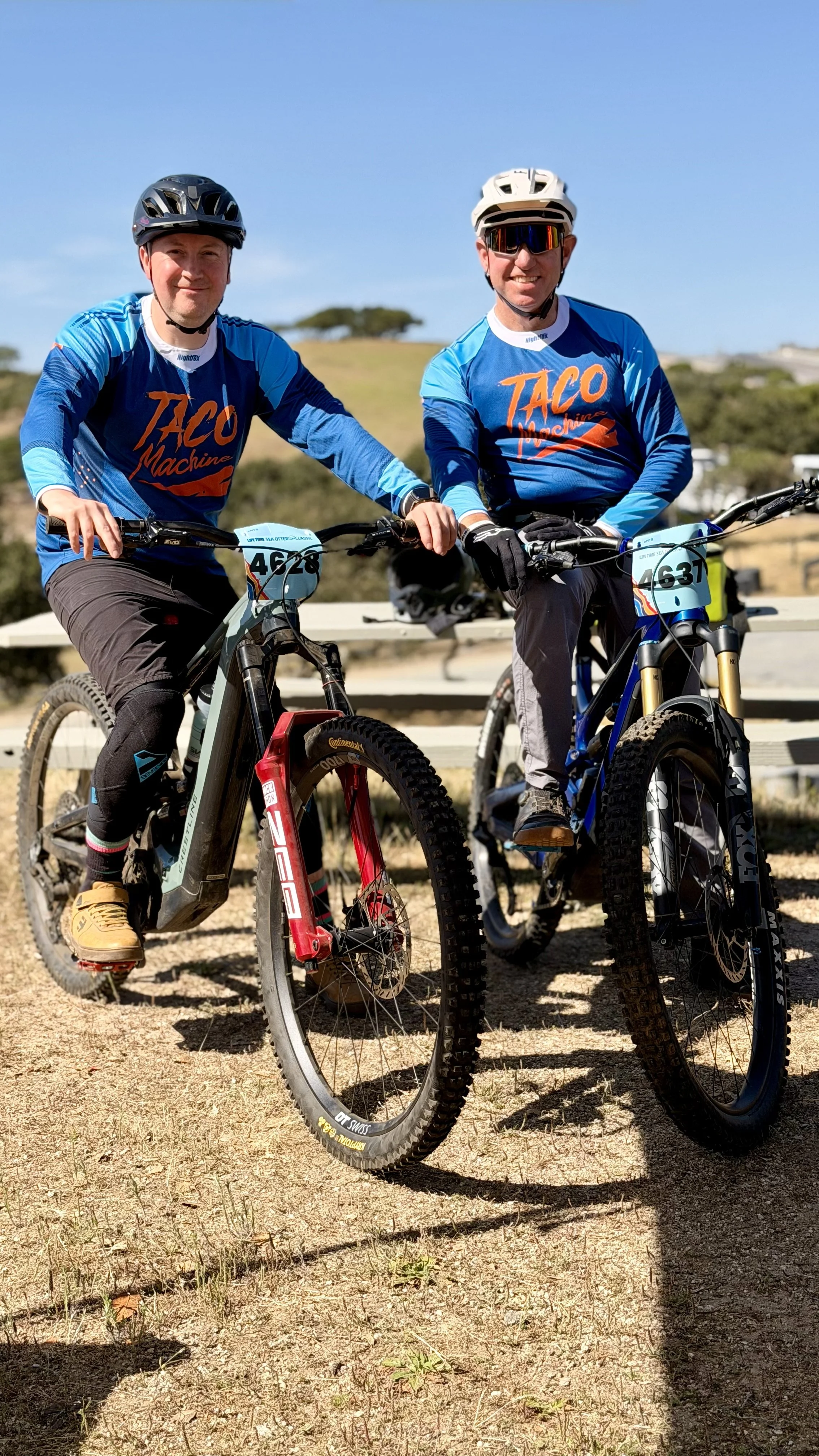 Two men wearing blue shirts, black pants, and helmets riding mountain bikes outdoors on a sunny day. They are smiling and looking at the camera, with a grassy hill and blue sky in the background.
