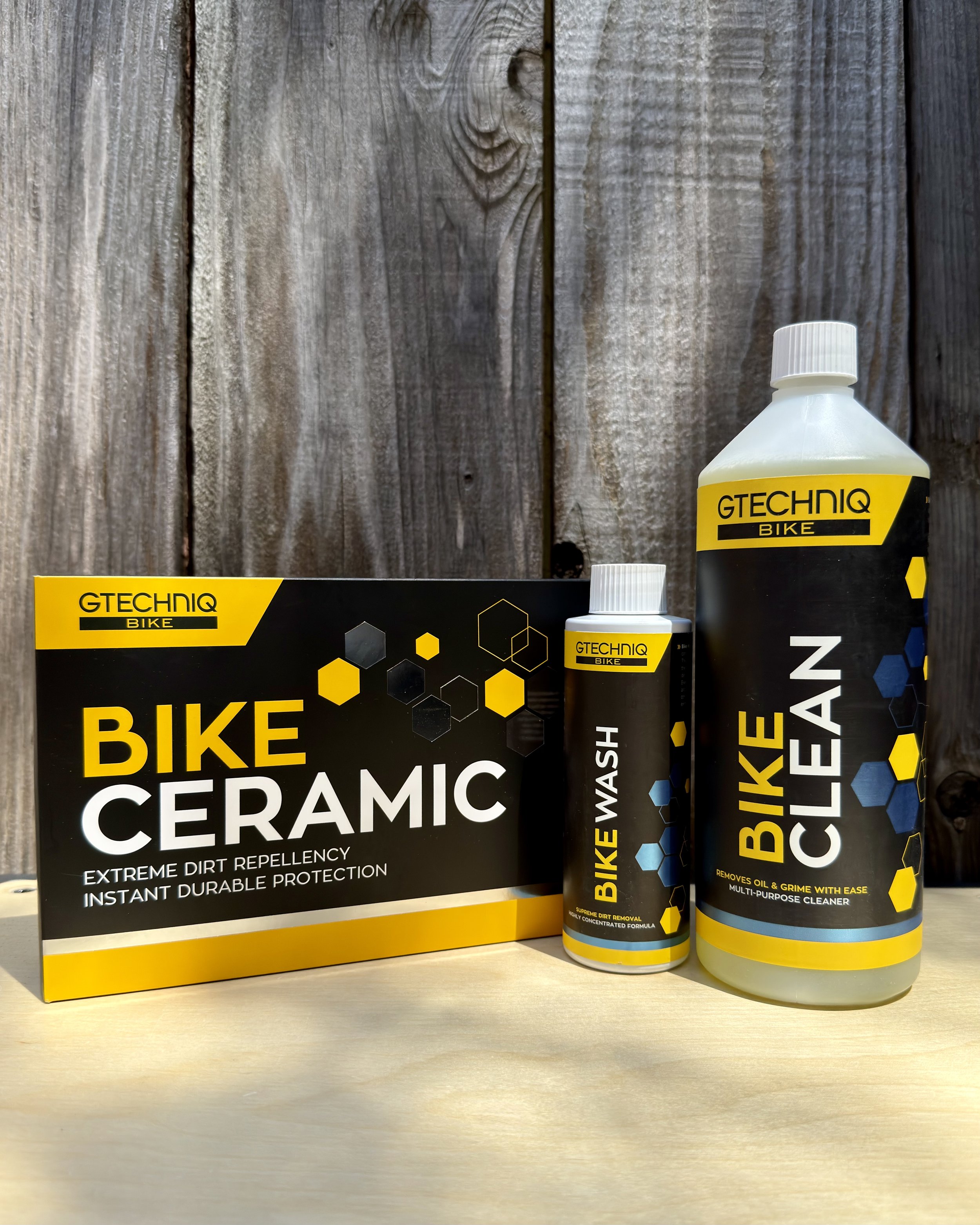 Two bottles of GTECHNIQ Bike Ceramic cleaner placed on a wooden surface with a black and yellow box beside them. The background shows a wooden wall.