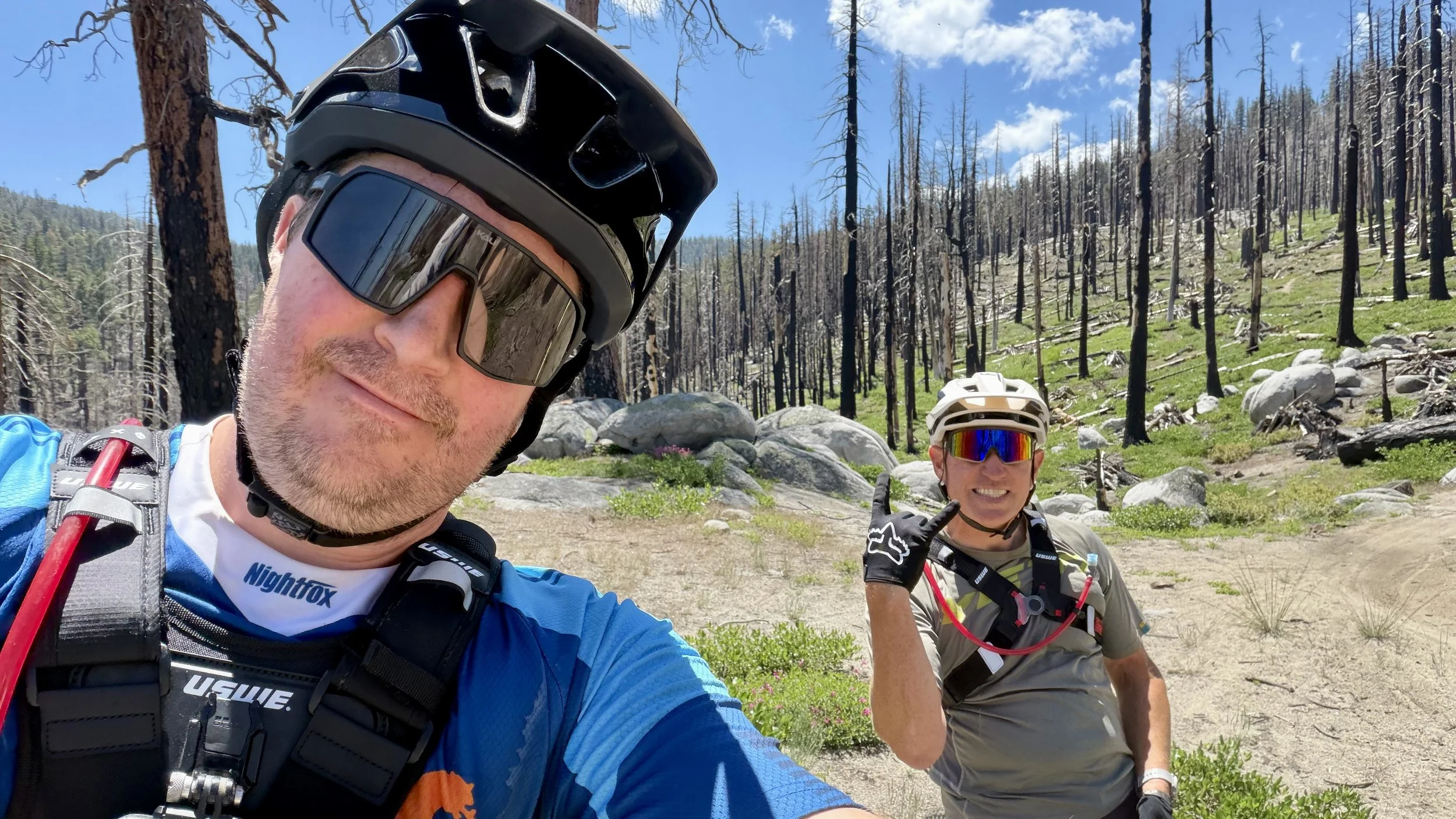 Two mountain bikers in protective gear taking a selfie outdoors in a forested area with rocks and trees, some trees charred from fire, under a partly cloudy sky.