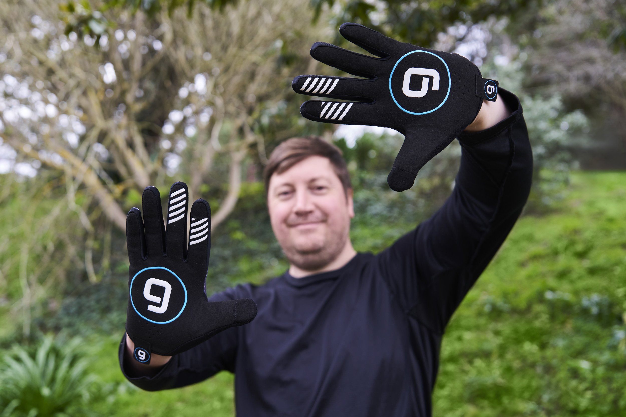 A man smiling outdoors, wearing black gloves with white and blue accents, holding up his hands in a gesture, with trees and greenery in the background.