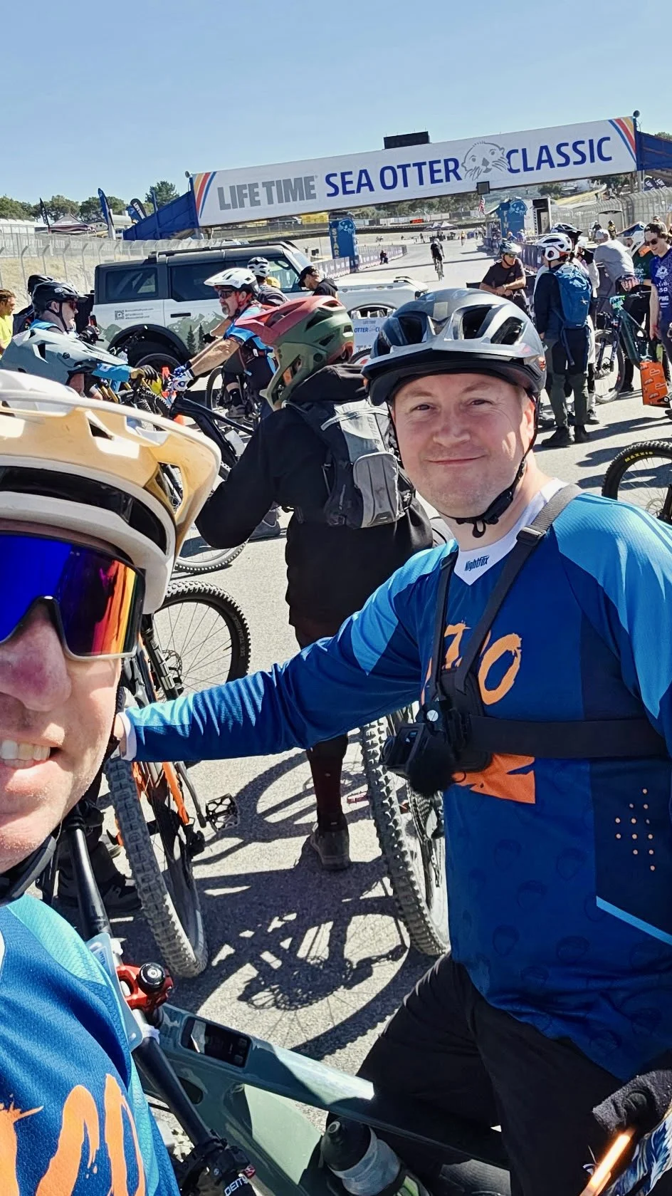 People in cycling gear gather at a start line under a sign reading 'LIFETIME SEA OTTER CLASSIC'. They are ready for a biking event outside on a sunny day.