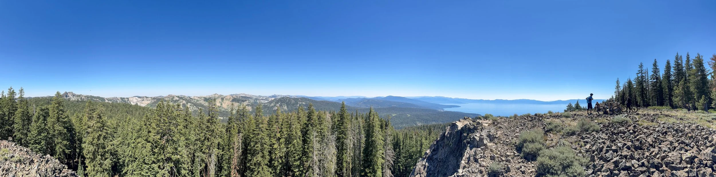 Panoramic view of a forested mountain landscape with a clear blue sky, rocky cliff edge on the right, and two people with bicycles standing near the edge overlooking the scene.