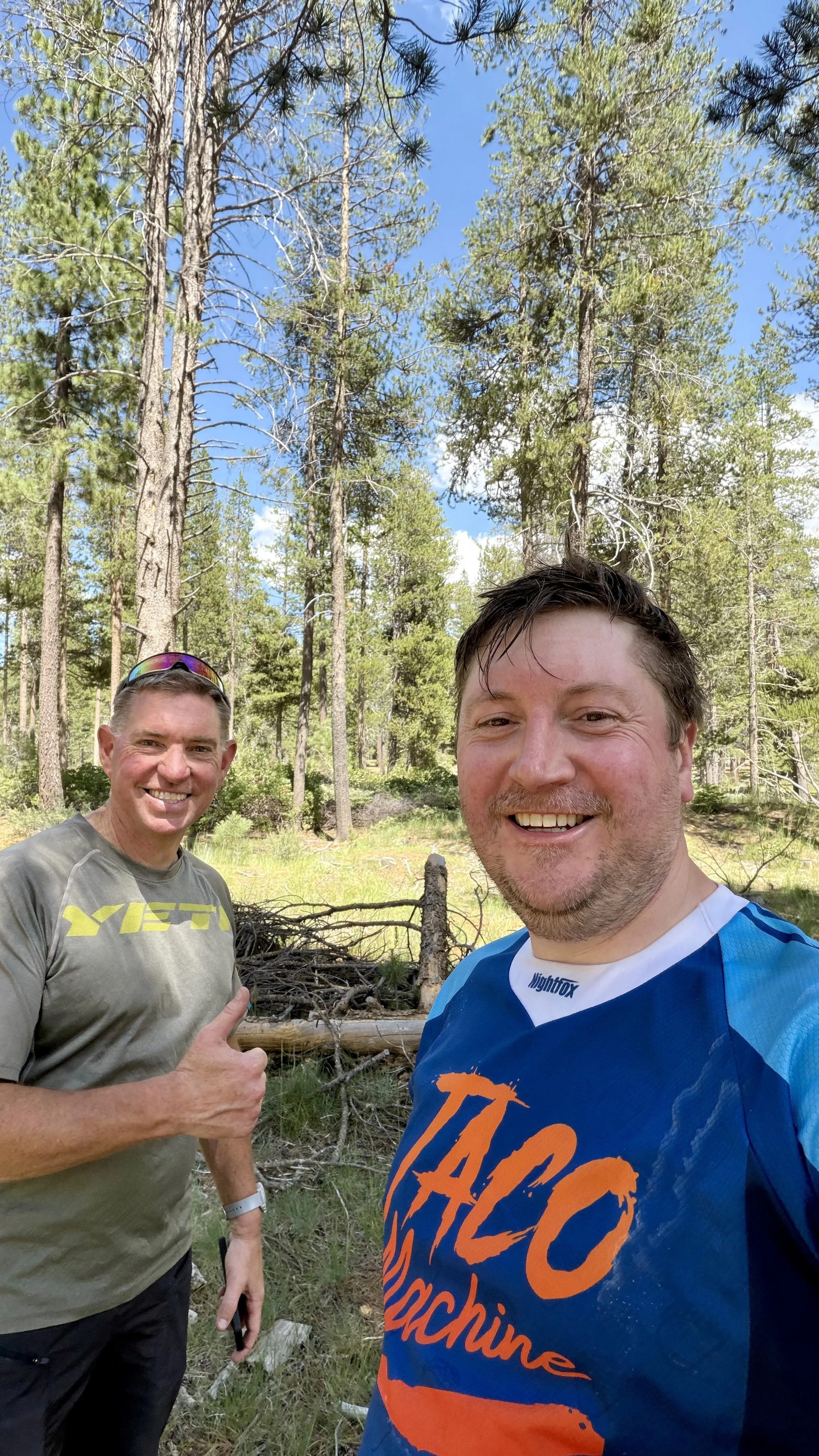 Two smiling men standing outdoors in a forest, surrounded by tall pine trees, with a blue sky overhead. One man is wearing a gray T-shirt with yellow lettering and sunglasses on his head, giving a thumbs-up. The other man is wearing a blue and orange