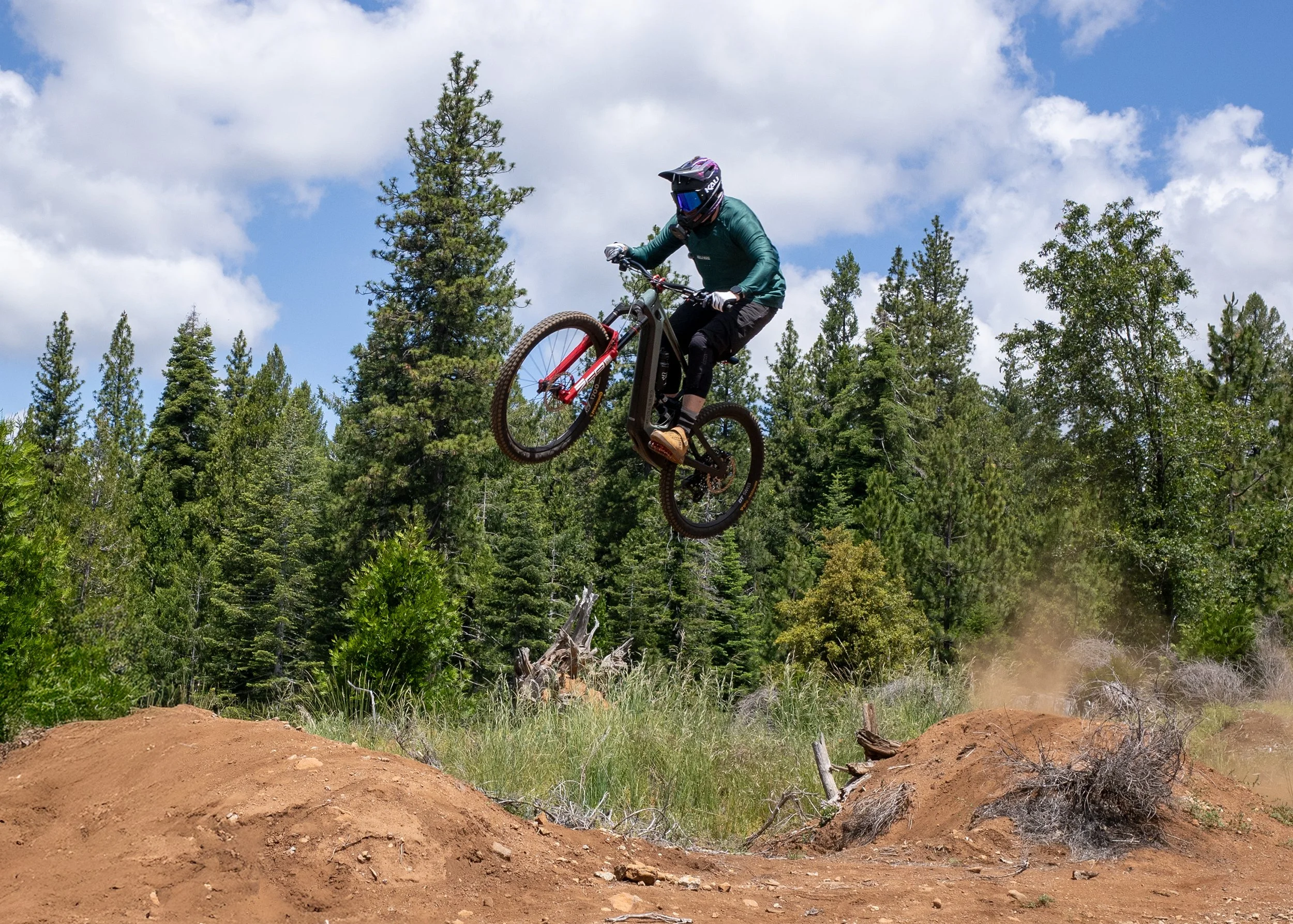 A mountain biker wearing a helmet and goggles is mid-air performing a jump over a dirt mound in a forested area with tall pine trees and a partly cloudy sky in the background.