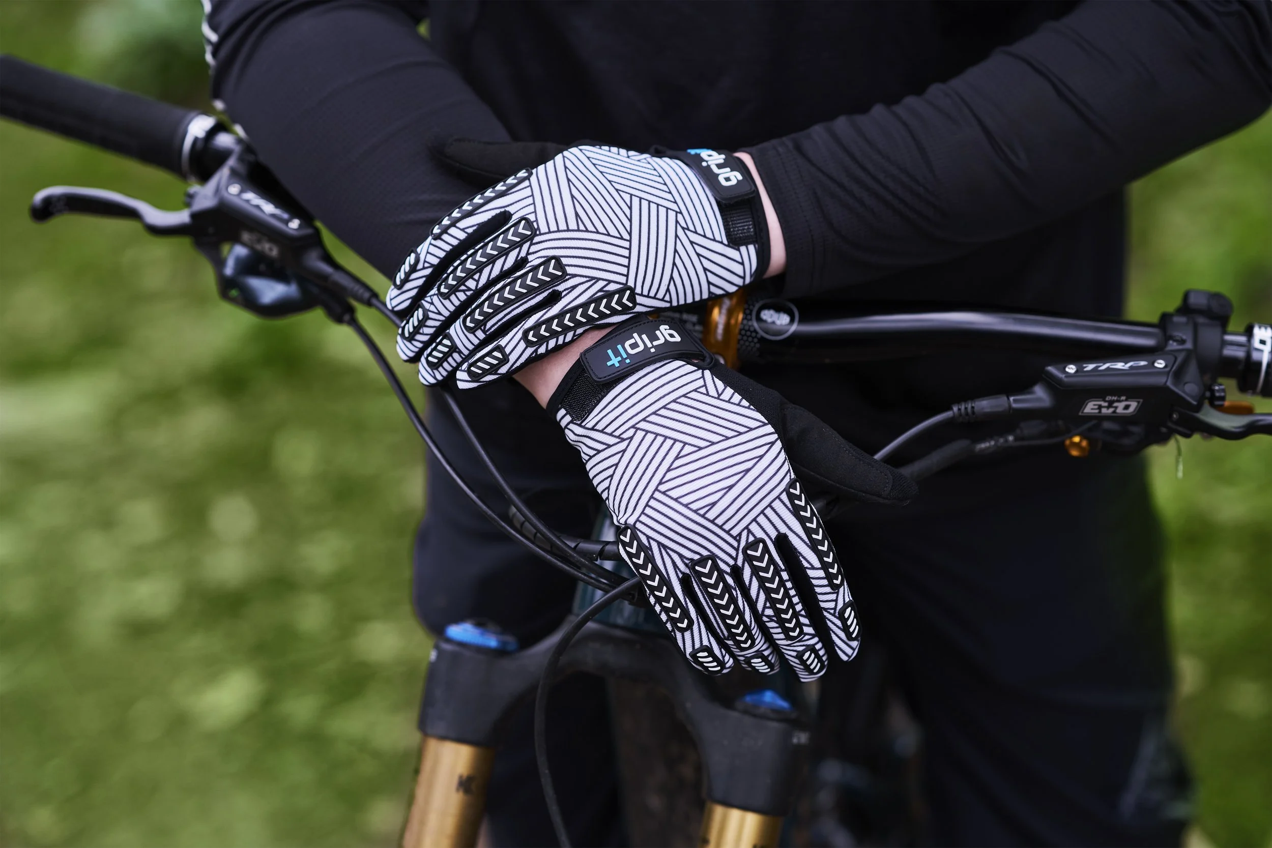 Close-up of a person wearing black gloves with white geometric patterns, gripping the handlebar of a mountain bike outdoors on grass.