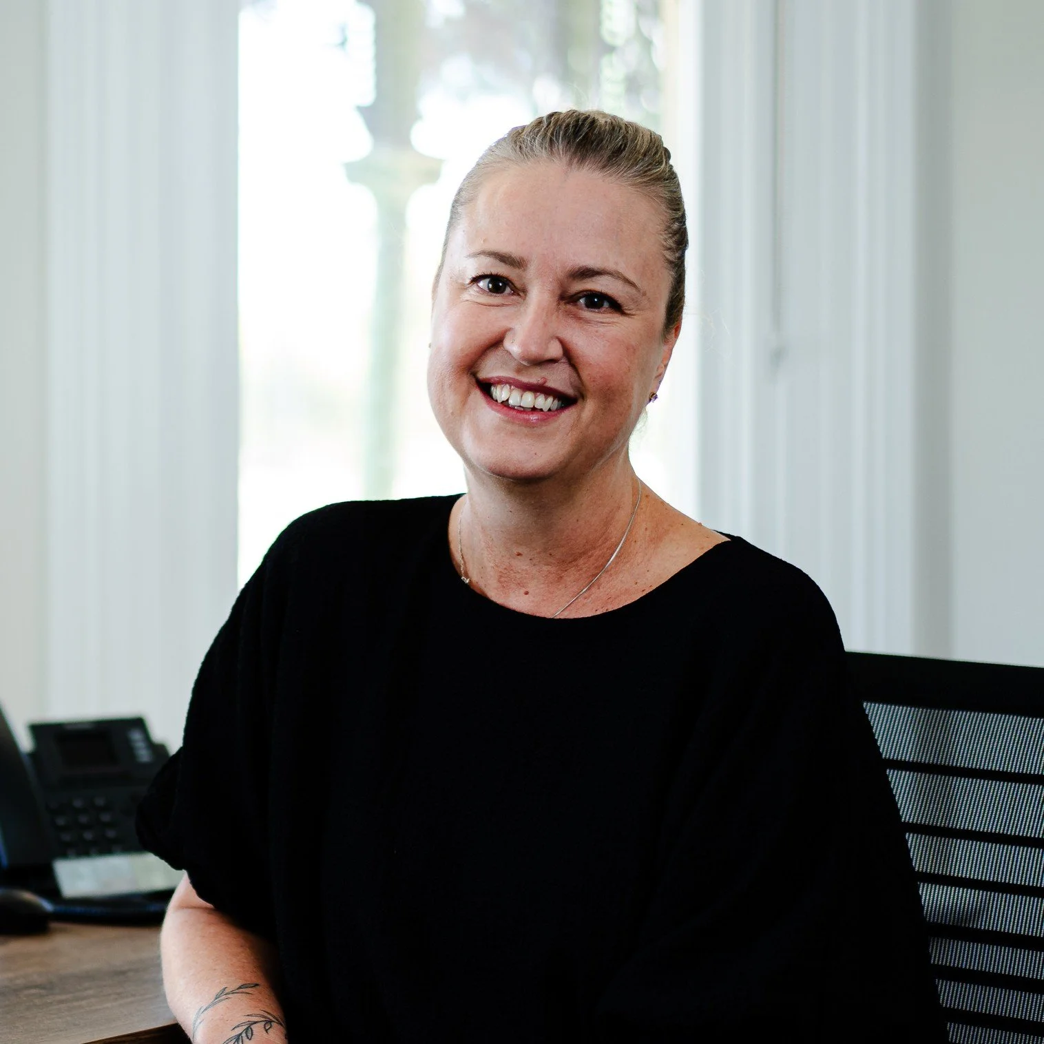 A woman smiling, sitting at a desk in an office with windows in the background.
