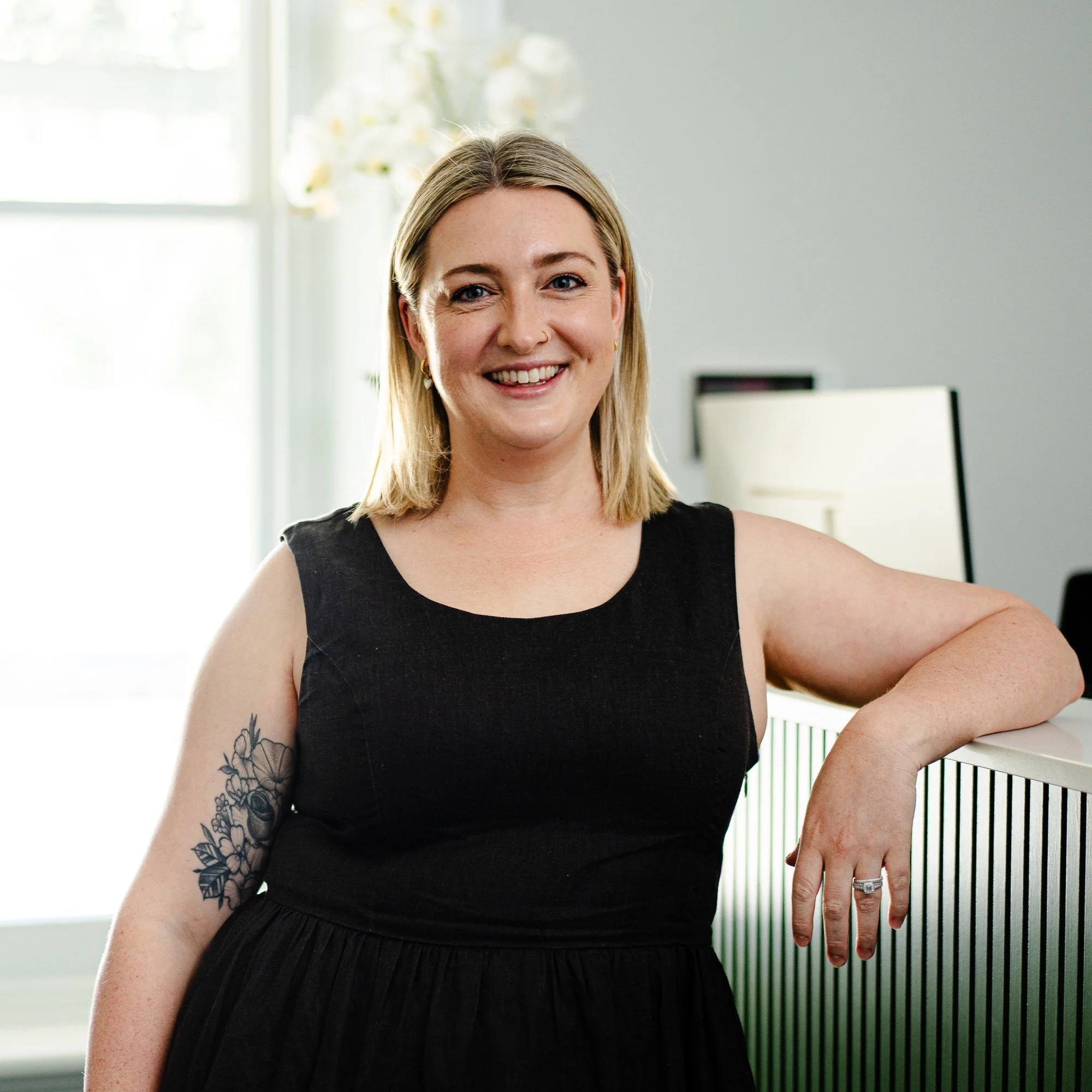 A woman with blonde hair, tattoos, and earrings smiling while standing outdoors, wearing a black sleeveless top, with her left arm resting on a railing.