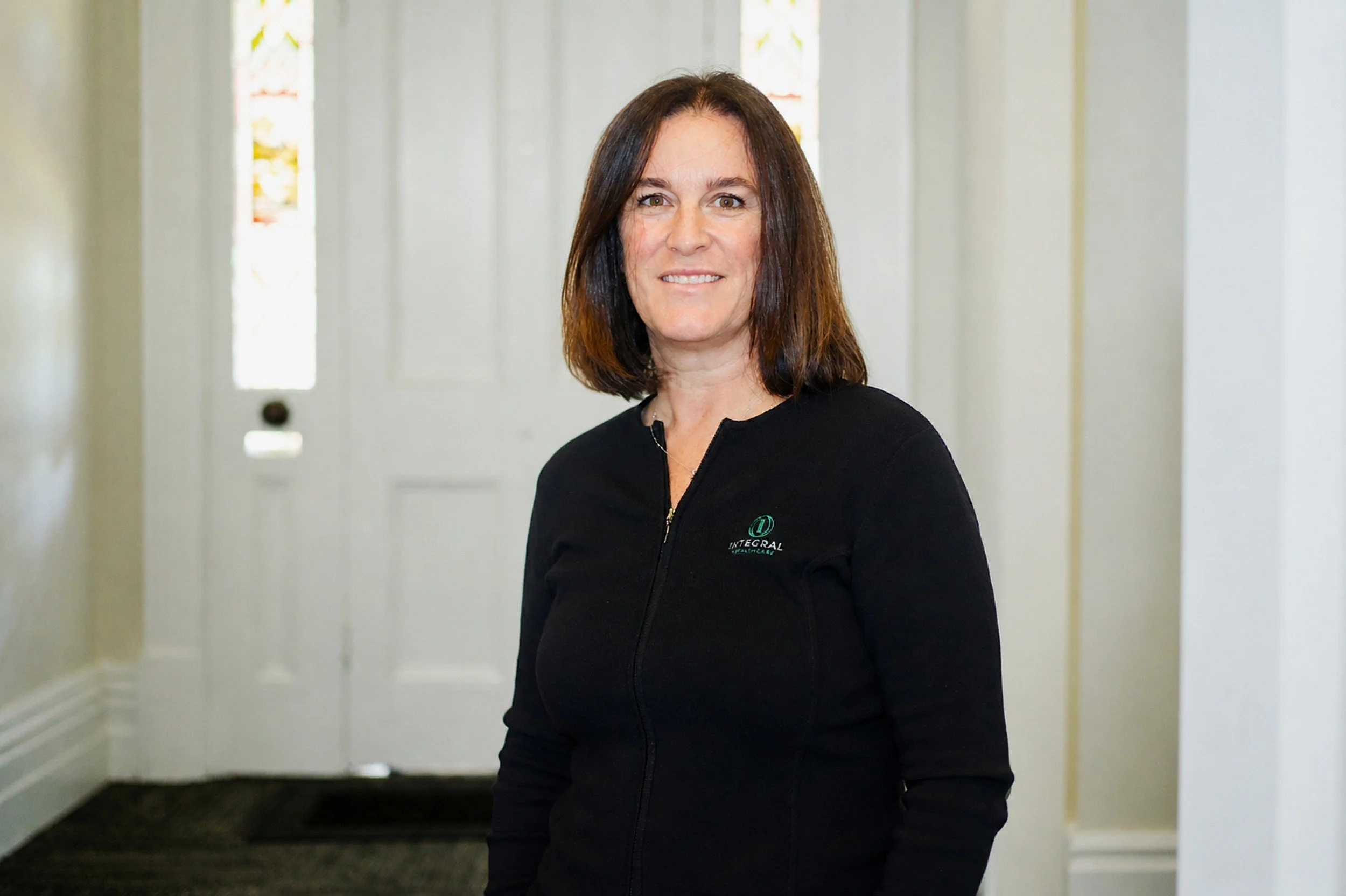 A woman with shoulder-length brown hair smiling at the camera, wearing a black zip-up jacket with a logo that says 'INTEGRAL' in a hallway with white doors and windows in the background.