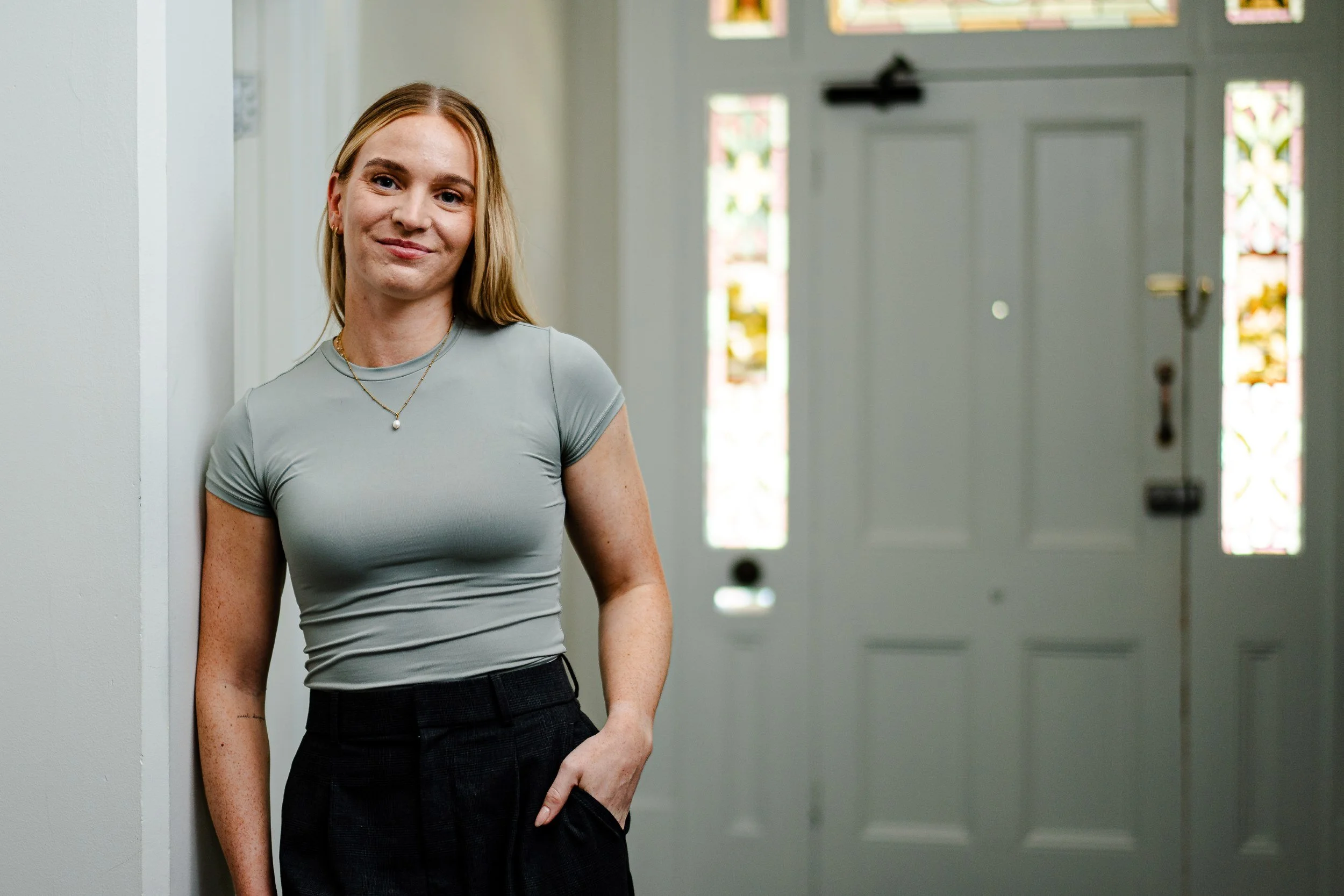 A woman with blonde hair, wearing a light gray t-shirt and black pants, leaning against a wall near a door with stained glass windows, smiling softly.