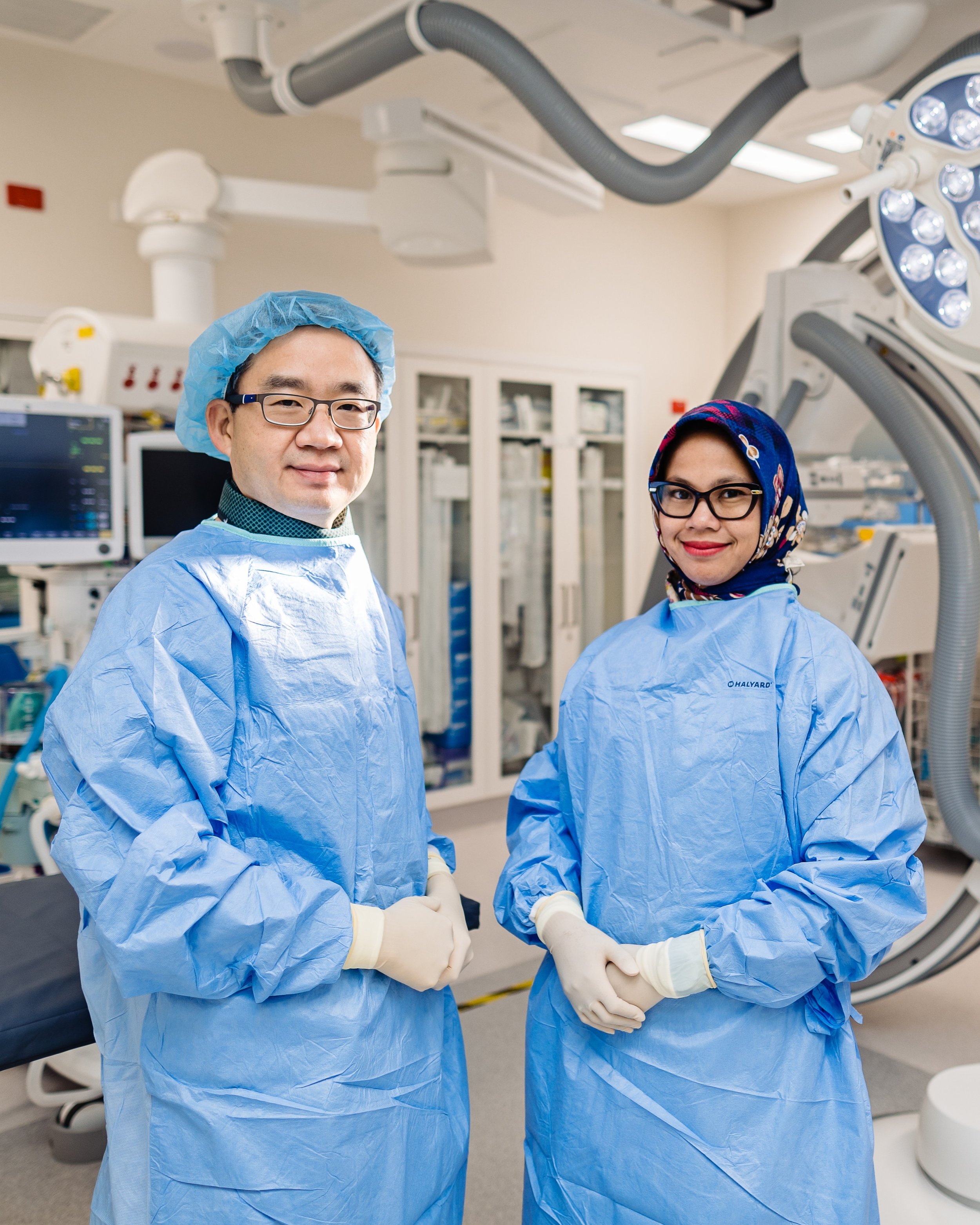 Two healthcare professionals in full surgical attire, including blue gowns, gloves, and head coverings, standing in a modern operating room with medical equipment and surgical lights overhead.