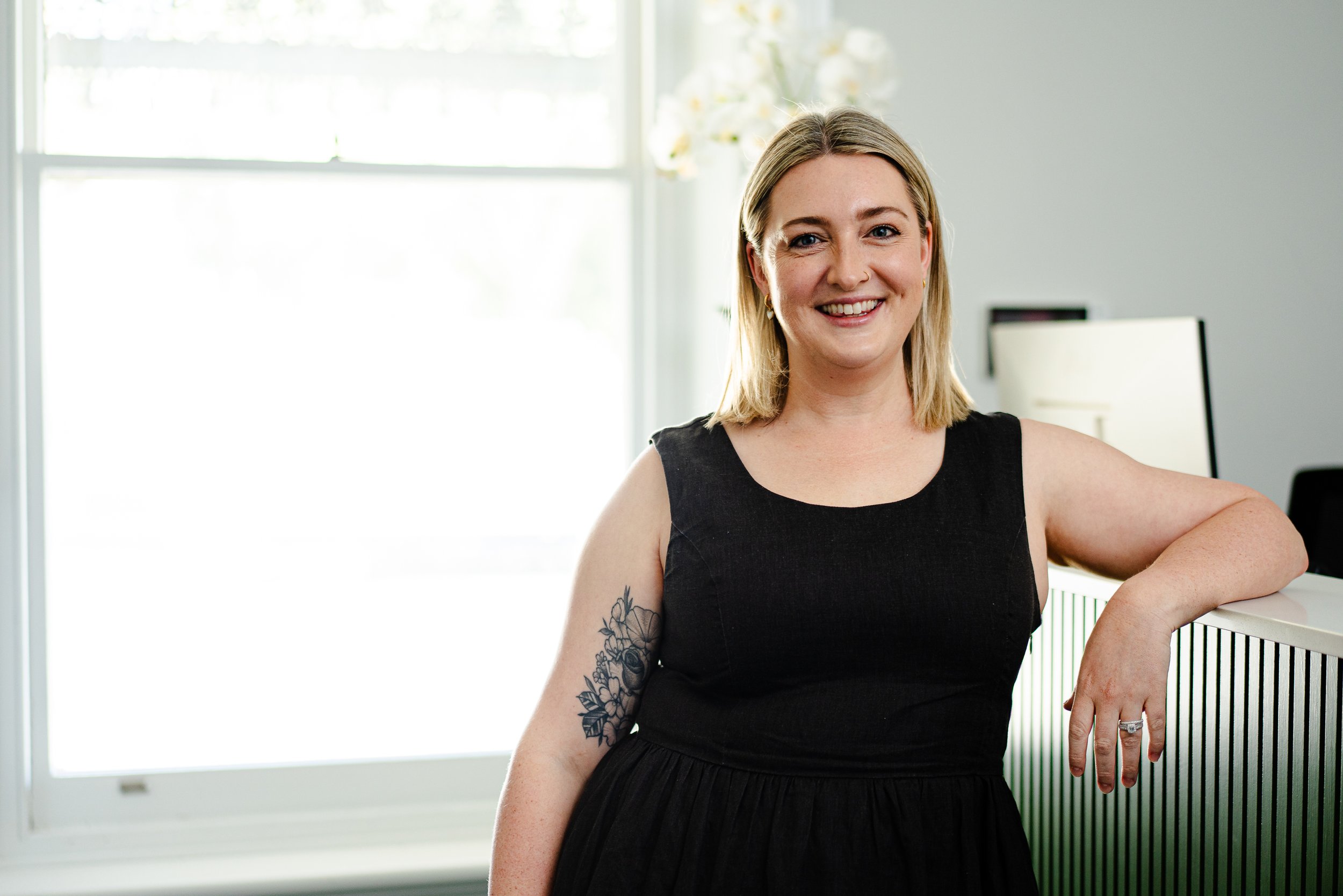 A smiling woman with shoulder-length blonde hair wearing a black sleeveless dress, standing near a white railing in a bright room with a large window and white flowers in the background.