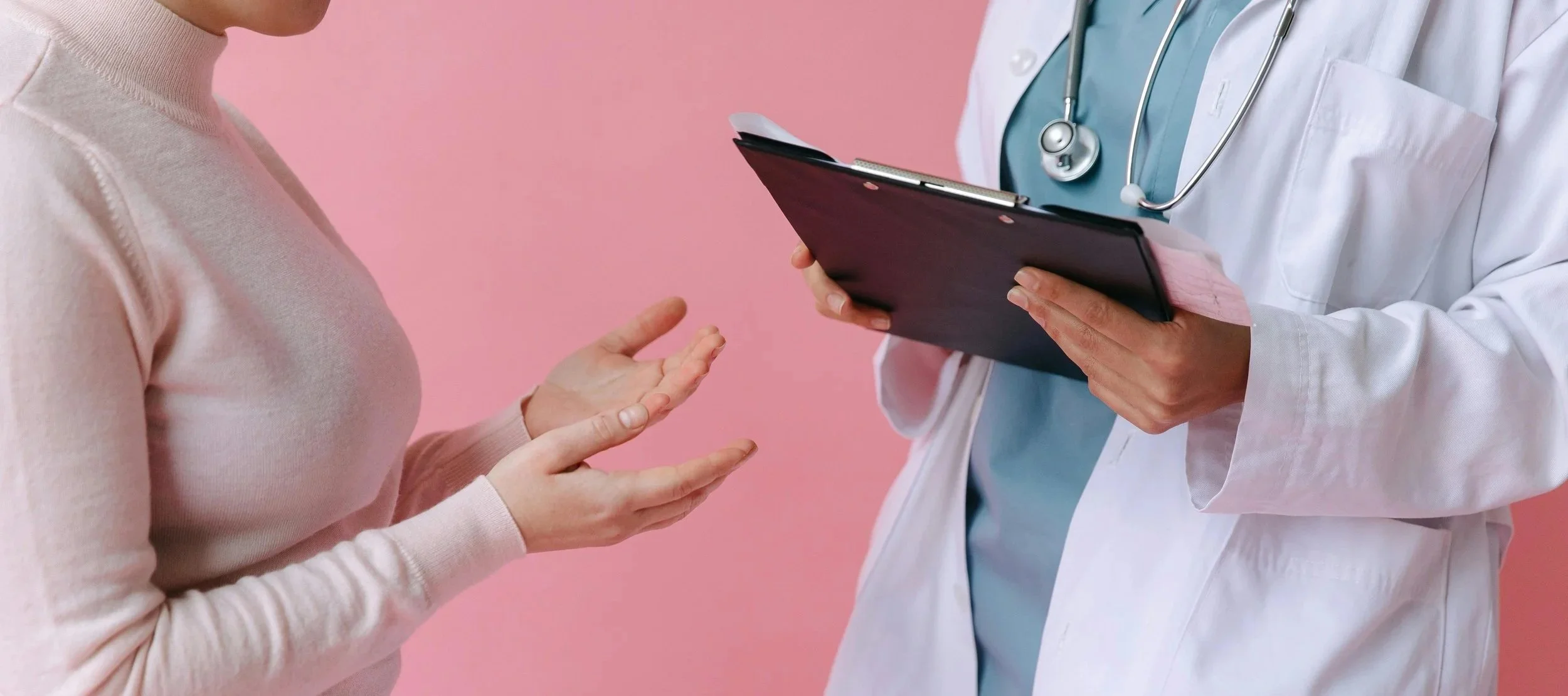 A healthcare professional in a white coat with a stethoscope around their neck shows a clipboard to a woman. The woman gestures with her hands during a consultation against a pink background.