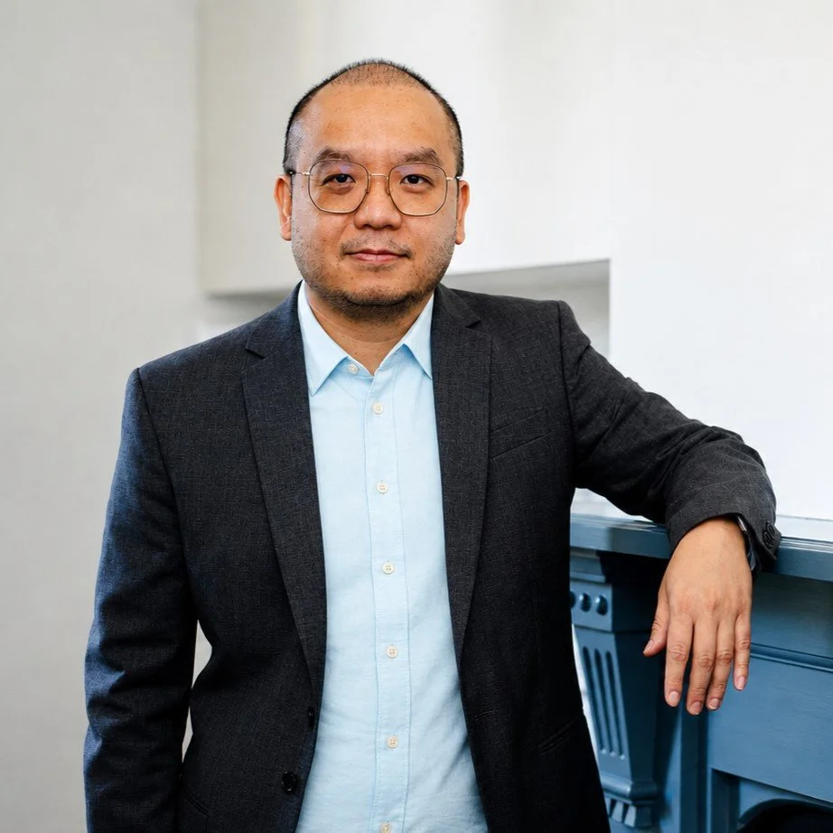 A man in a dark blazer and light blue shirt with glasses, standing with his right arm resting on a blue upright piano in a minimalistic, white-walled room.