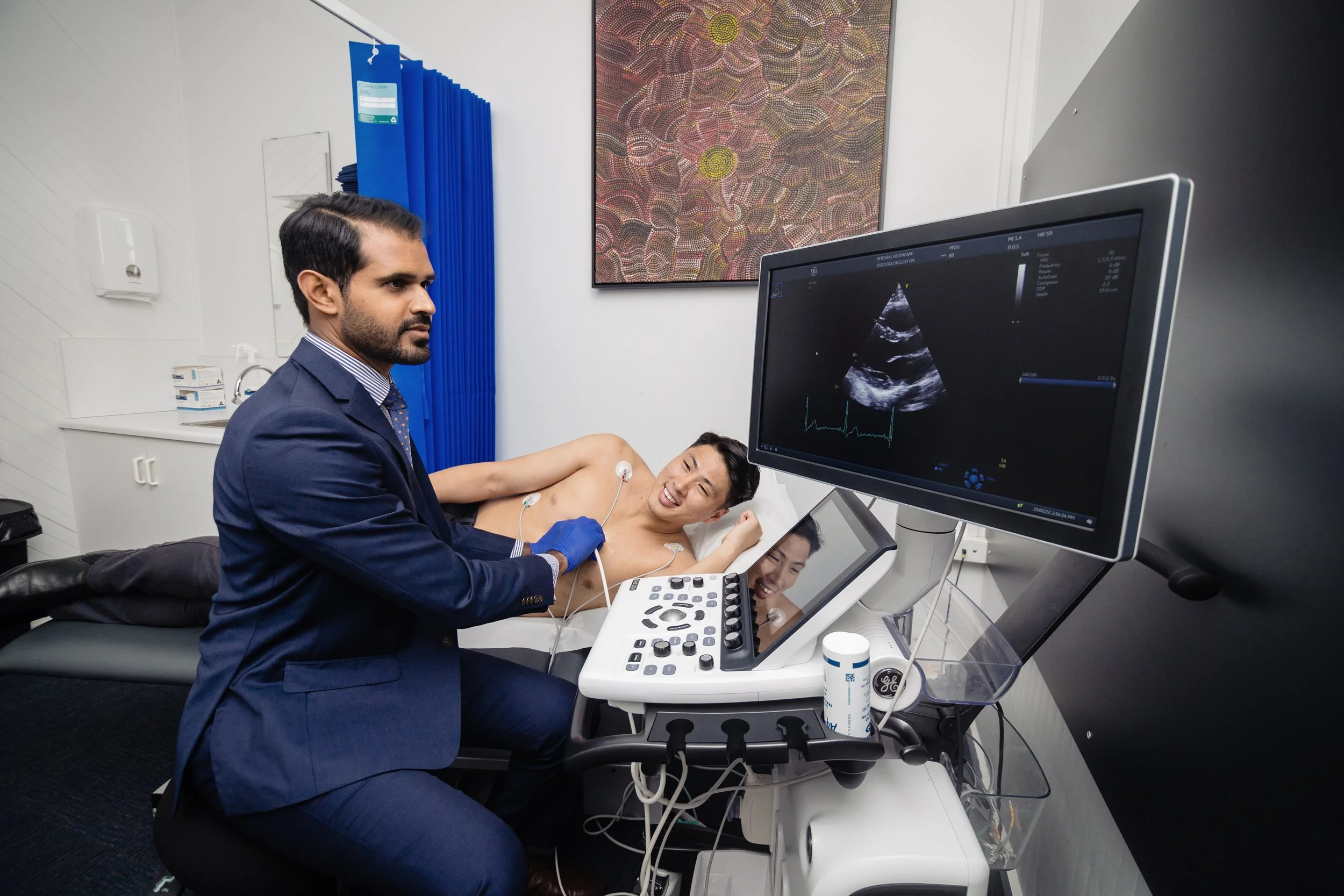 Doctor performing an ultrasound procedure on a female patient lying on a hospital bed, with ultrasound images visible on a monitor.