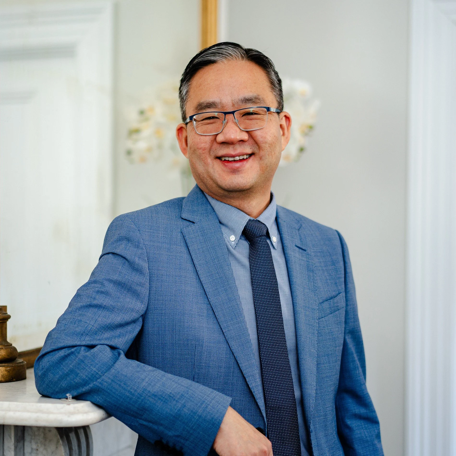 A man in a blue suit and glasses smiling, leaning against a marble countertop in a bright room.
