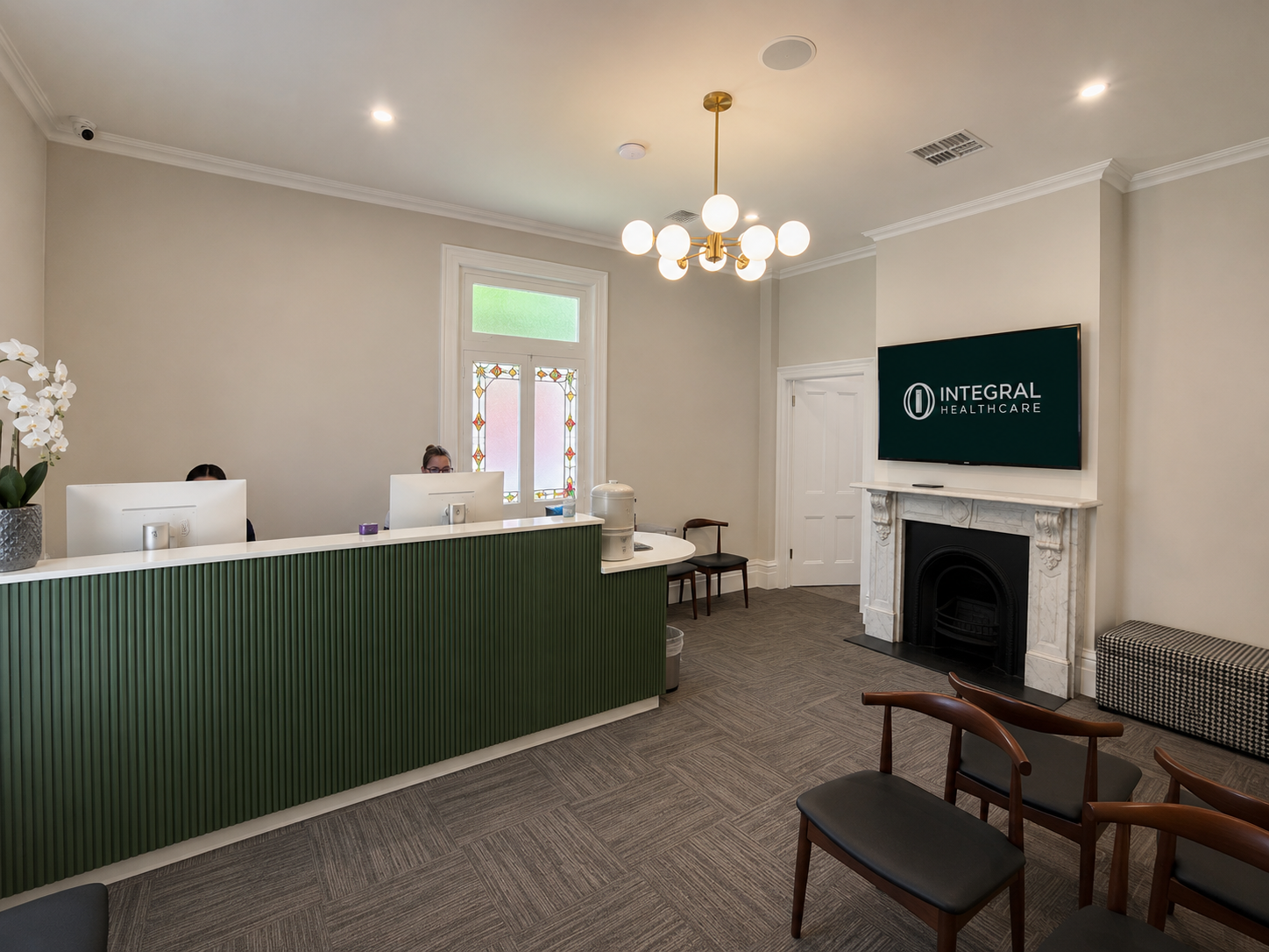 Reception area of a healthcare facility with a green front desk, two receptionists working on computers, a decorative window, a TV screen displaying 'Integral Healthcare', a fireplace, chairs, and a patterned bench.