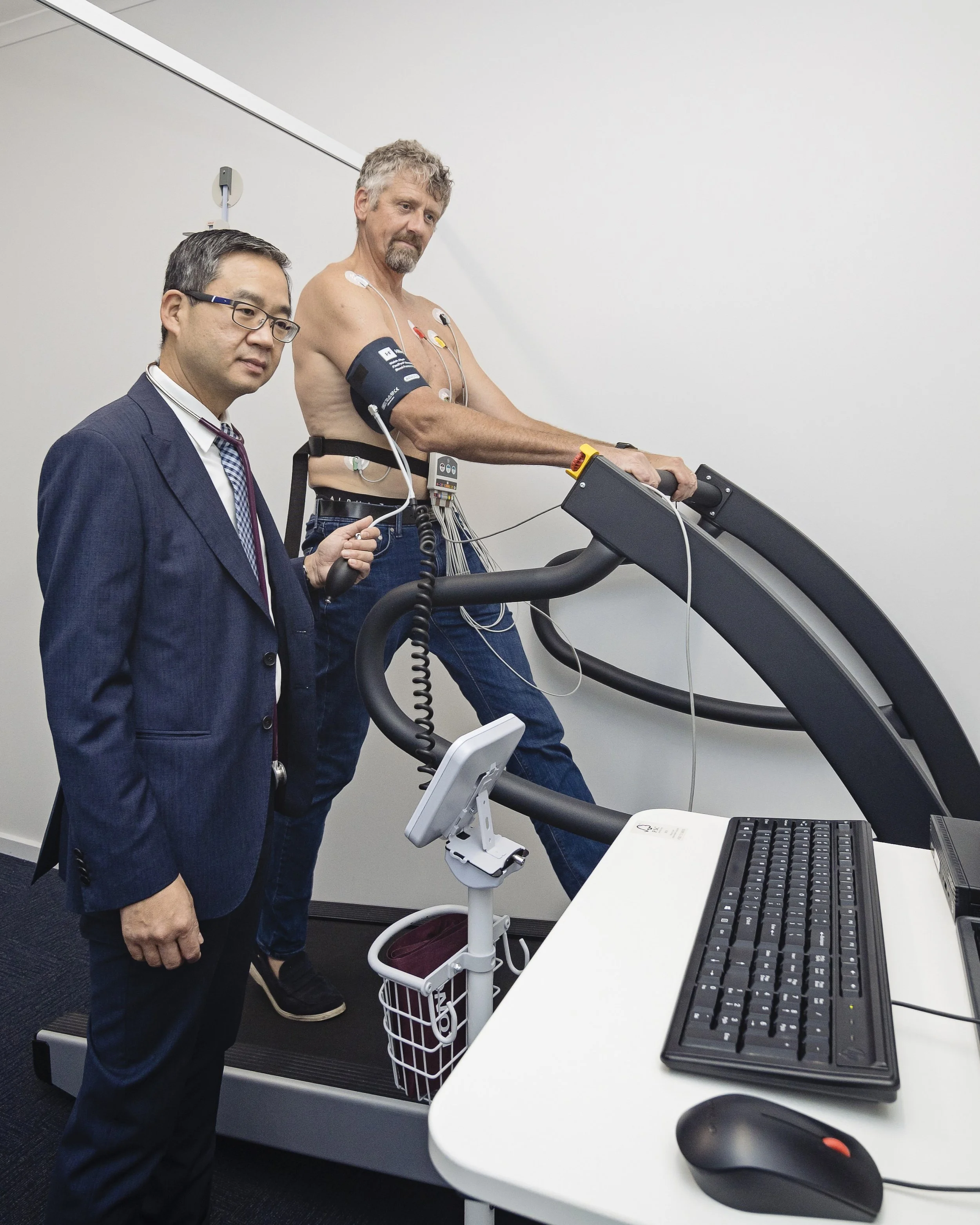 A man with medical electrodes on his chest and arm walking on a treadmill in a clinical room, with a doctor or technician supervising, and a computer keyboard and mouse on a white desk nearby.
