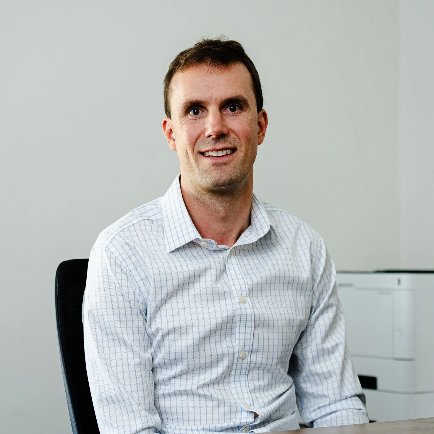 A man with short dark hair, wearing a light blue and white checkered dress shirt, sitting in a black office chair in an office setting.