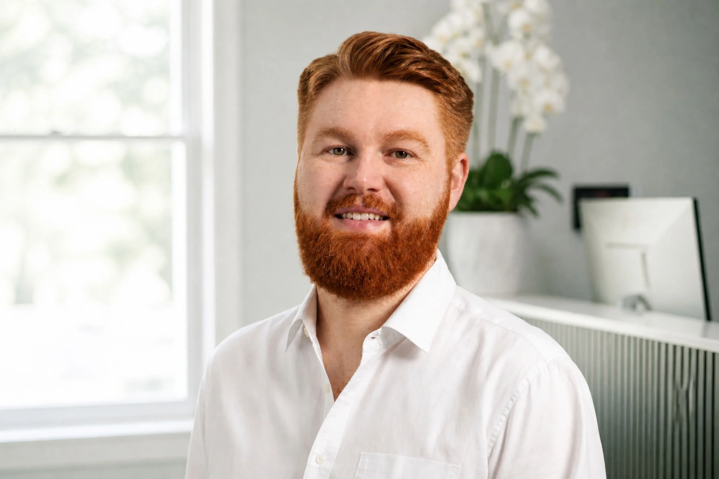 A man with red hair and a beard wearing a white shirt, standing in a bright room with large windows and a potted plant in the background.