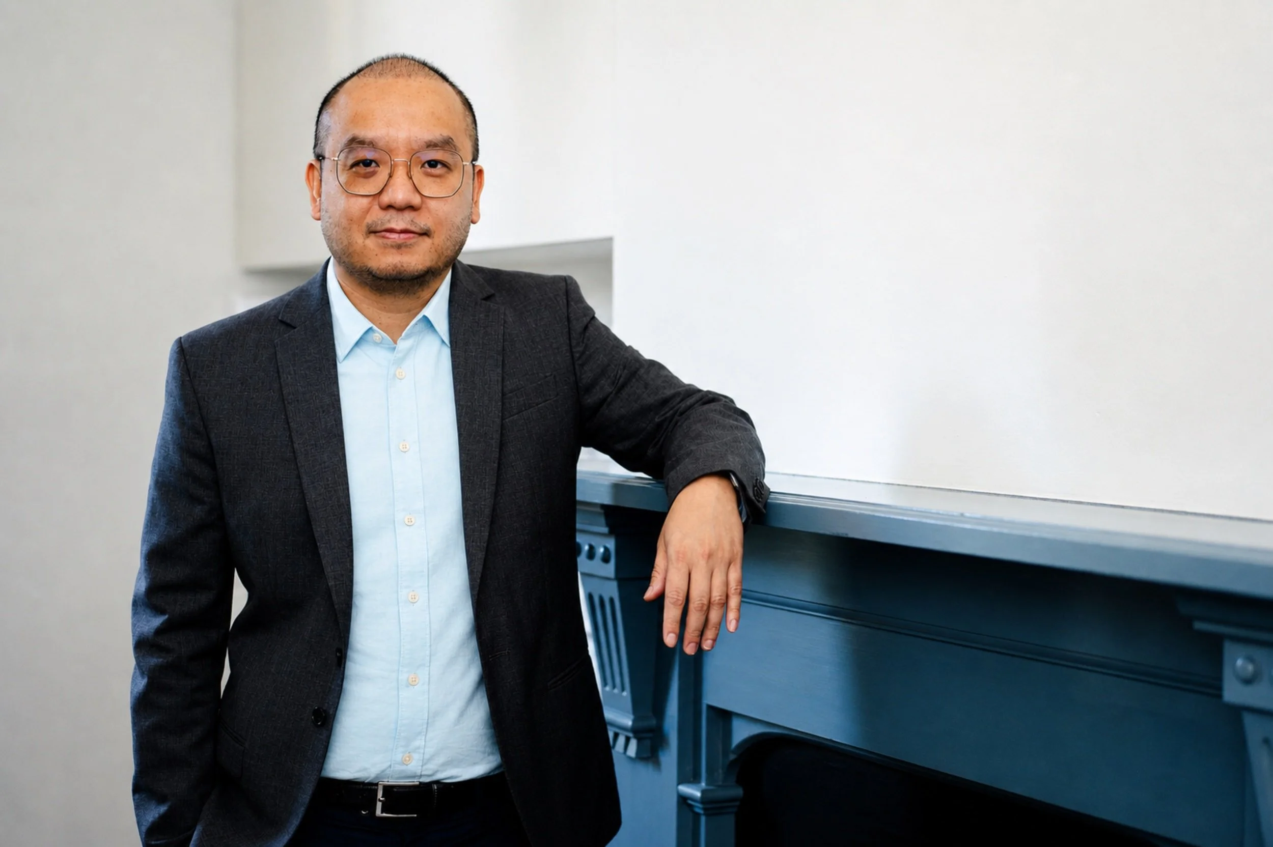 A man with glasses in a suit jacket and light blue shirt standing next to a blue piano in a room with plain white walls.