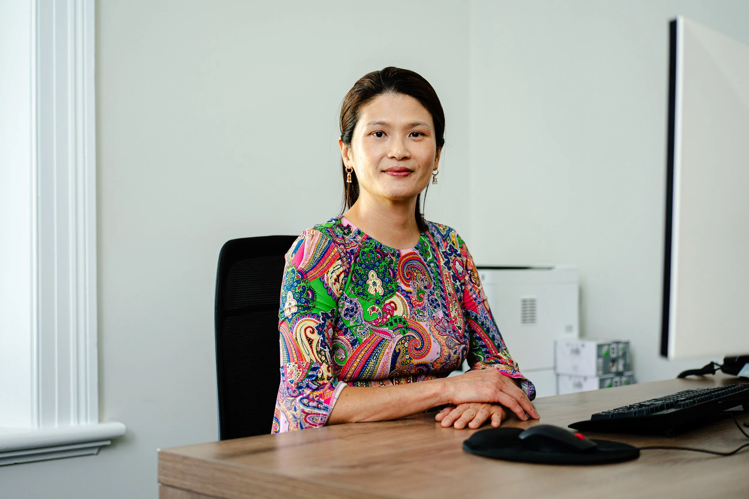 A woman with dark hair sitting at a desk in an office, wearing a colorful, paisley-patterned blouse, with a computer and keyboard in front of her.
