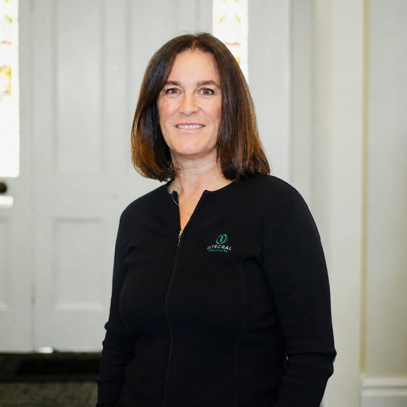 A woman with shoulder-length brown hair, wearing a black zip-up jacket with a logo, standing in front of a white door with small decorative windows.