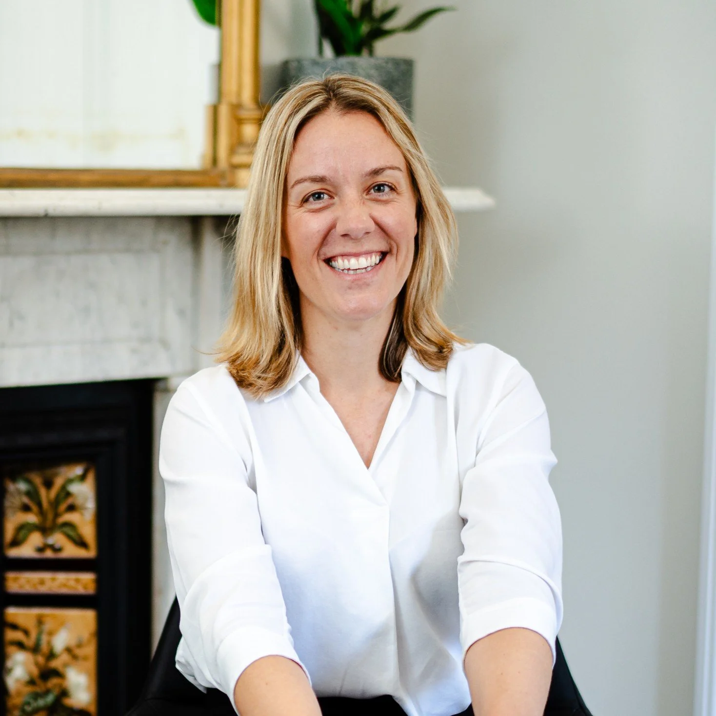 A smiling woman with shoulder-length blonde hair wearing a white blouse sitting indoors with background elements including a plant, a mirror, and decorative woodwork.
